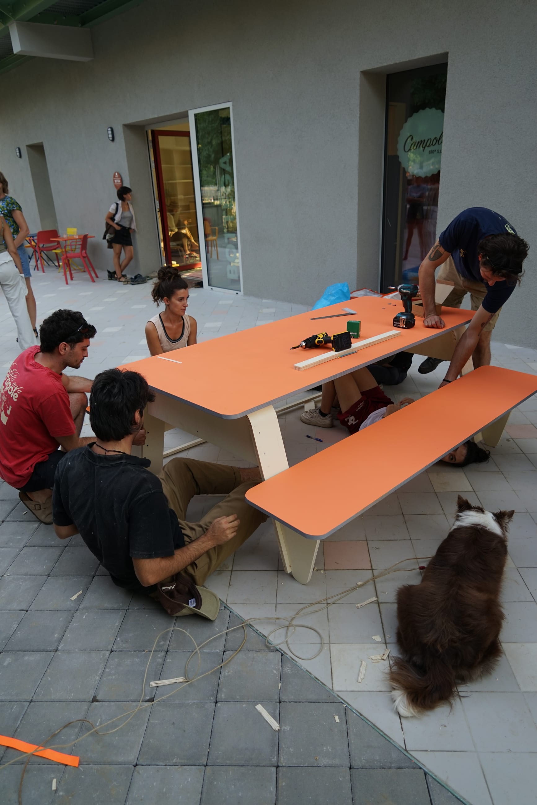 Persone montano un tavolo con panca arancione su una terrazza, accanto a un cane