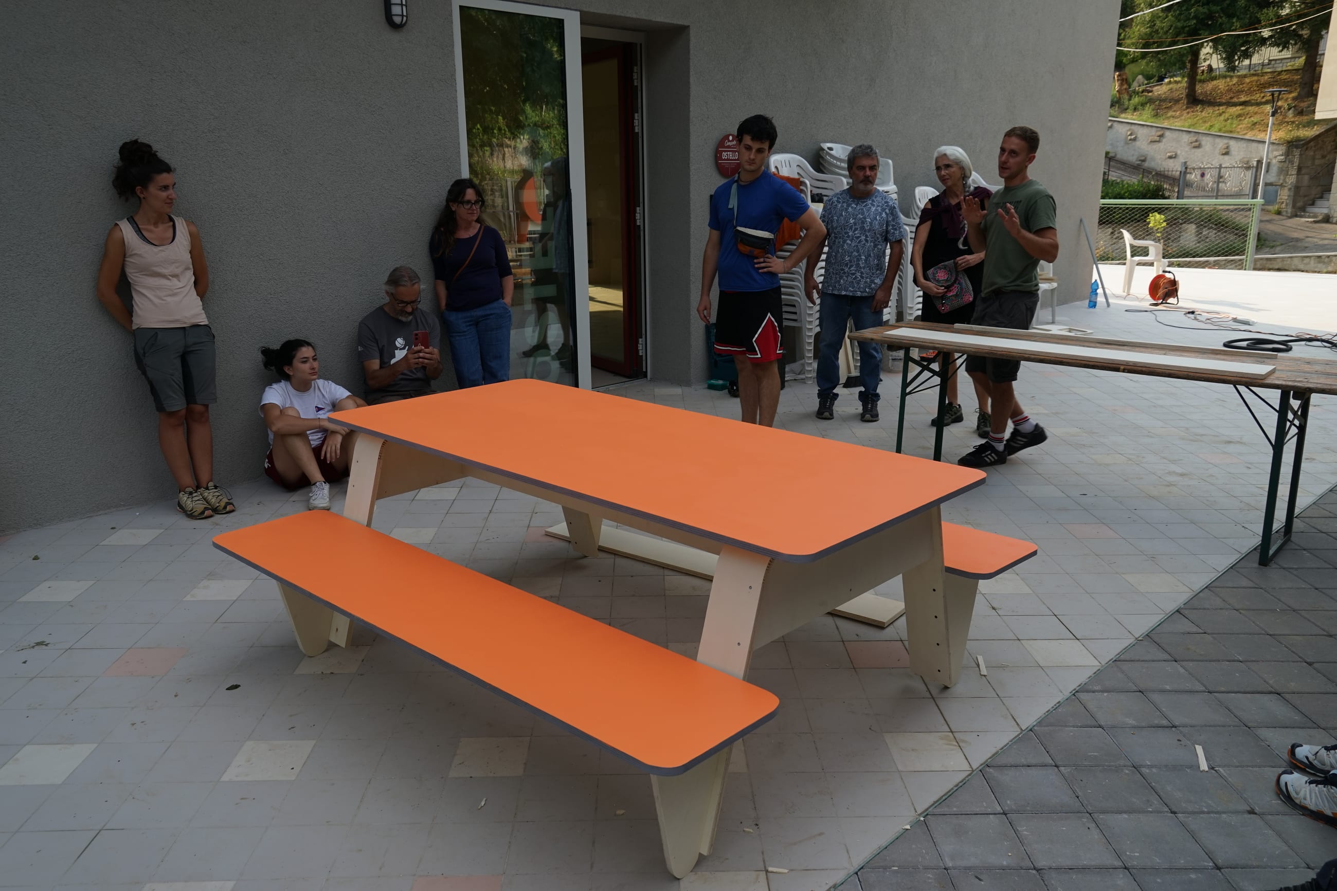Tavolo con panche in legno arancione su una terrazza, con persone in piedi attorno