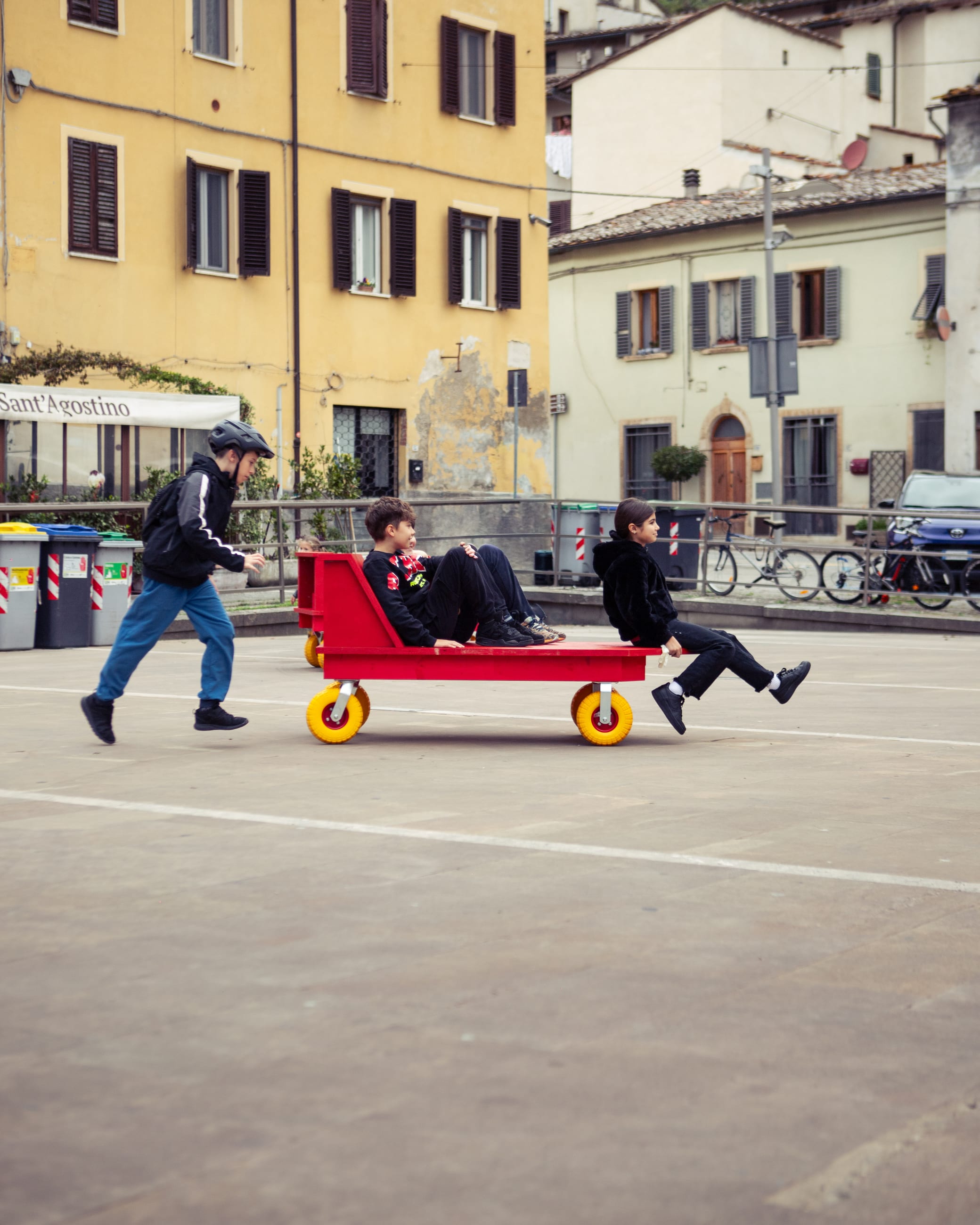 Due ragazzi su un carrello rosso a ruote gialle spinto da una persona in una piazza