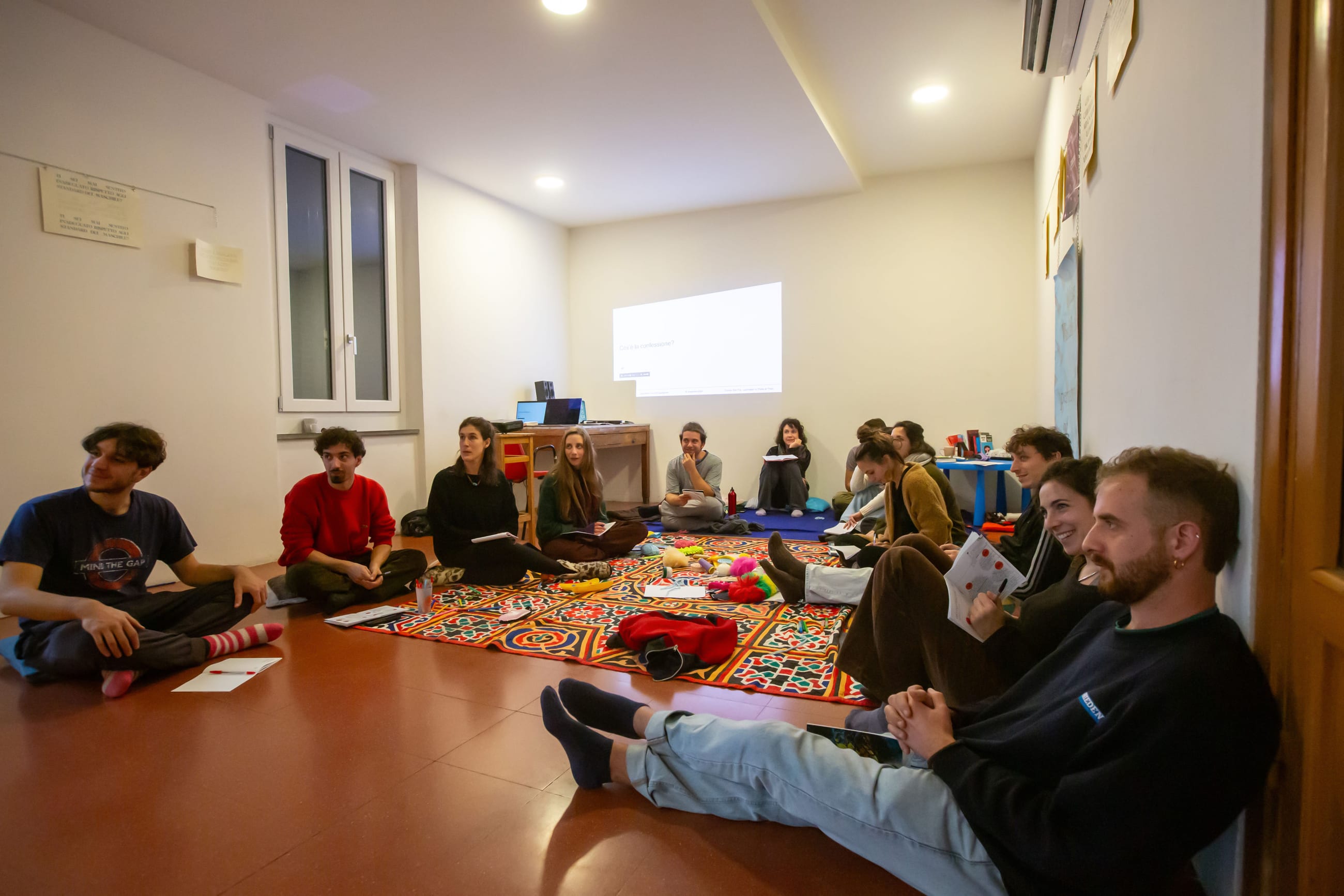 Persone sedute a terra in cerchio su tappeti colorati, in una stanza durante un incontro