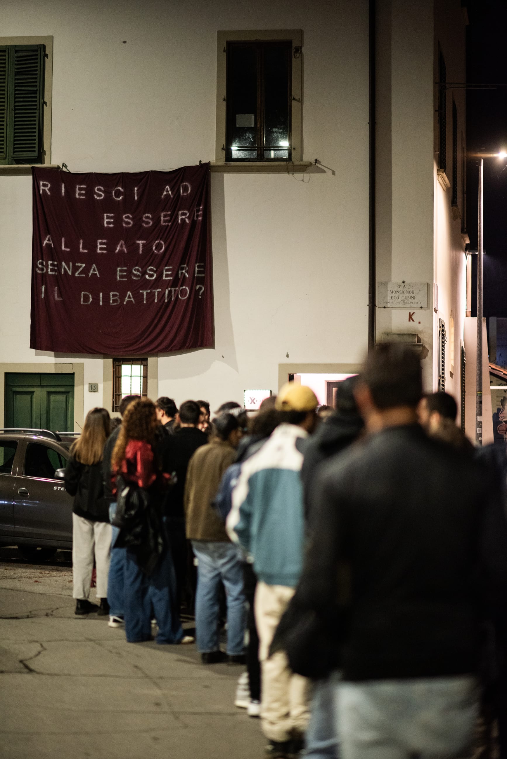 Persone in fila di sera davanti a un edificio con uno striscione appeso alla facciata