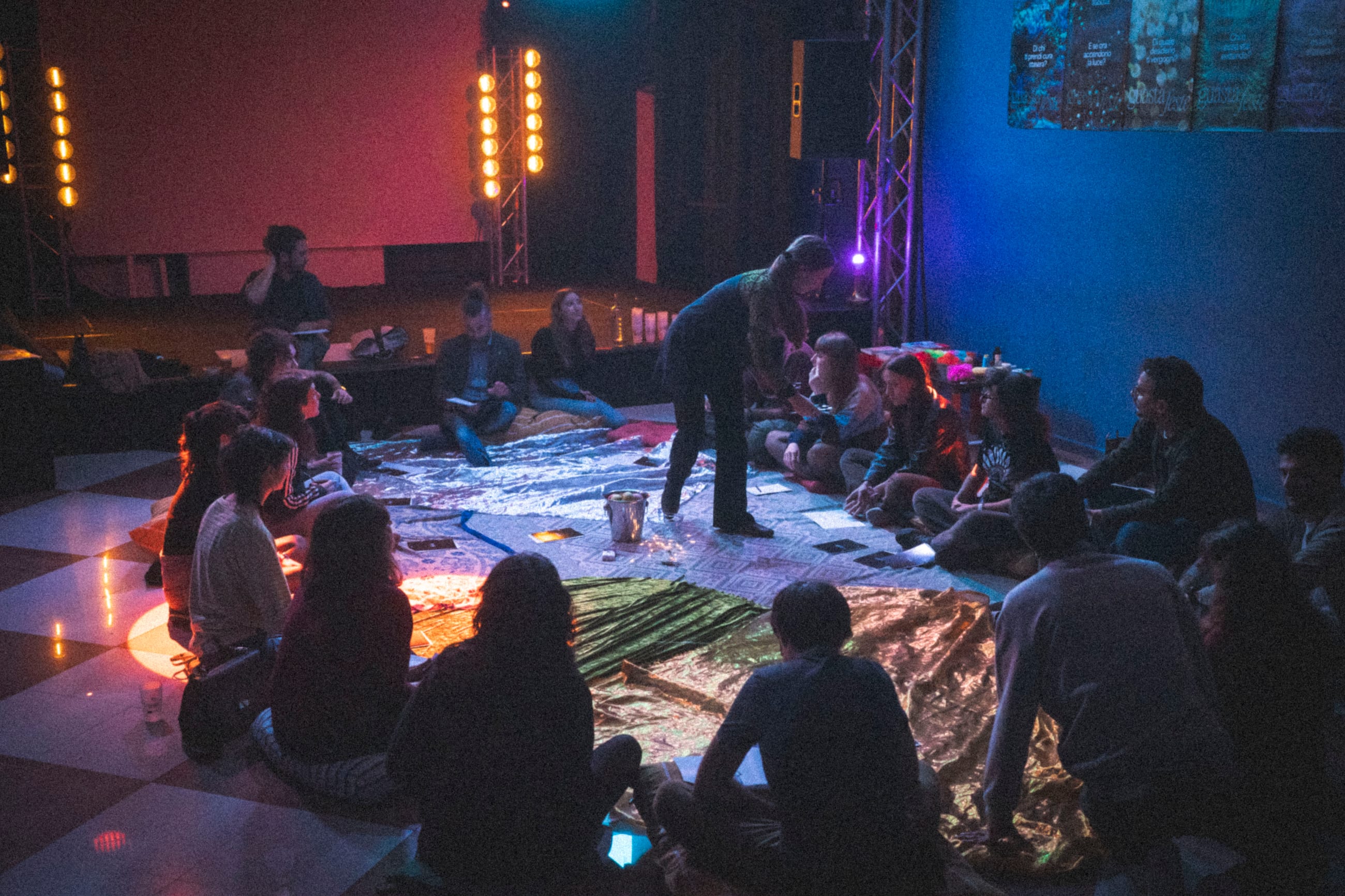 Persone sedute a terra su teli argentati in una sala buia con luci colorate
