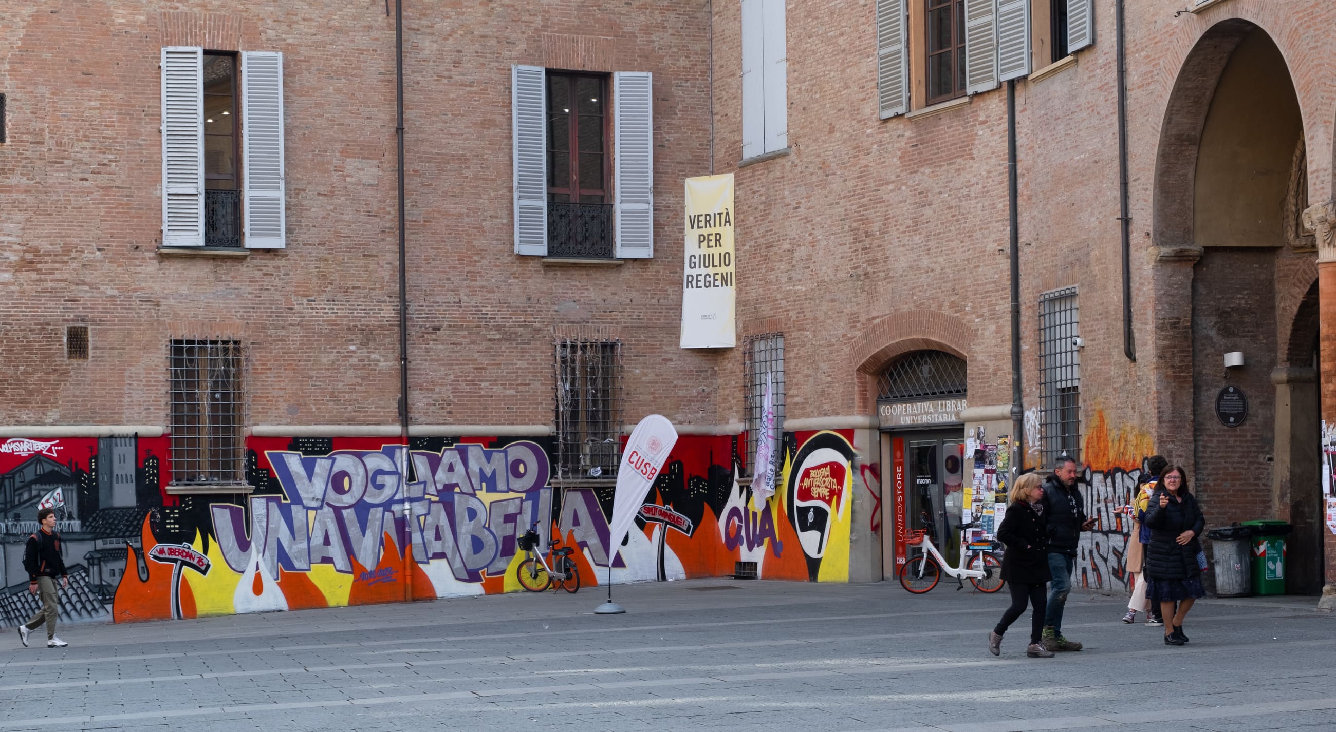Piazza con un edificio in mattoni e un lungo murale colorato, attraversata da persone