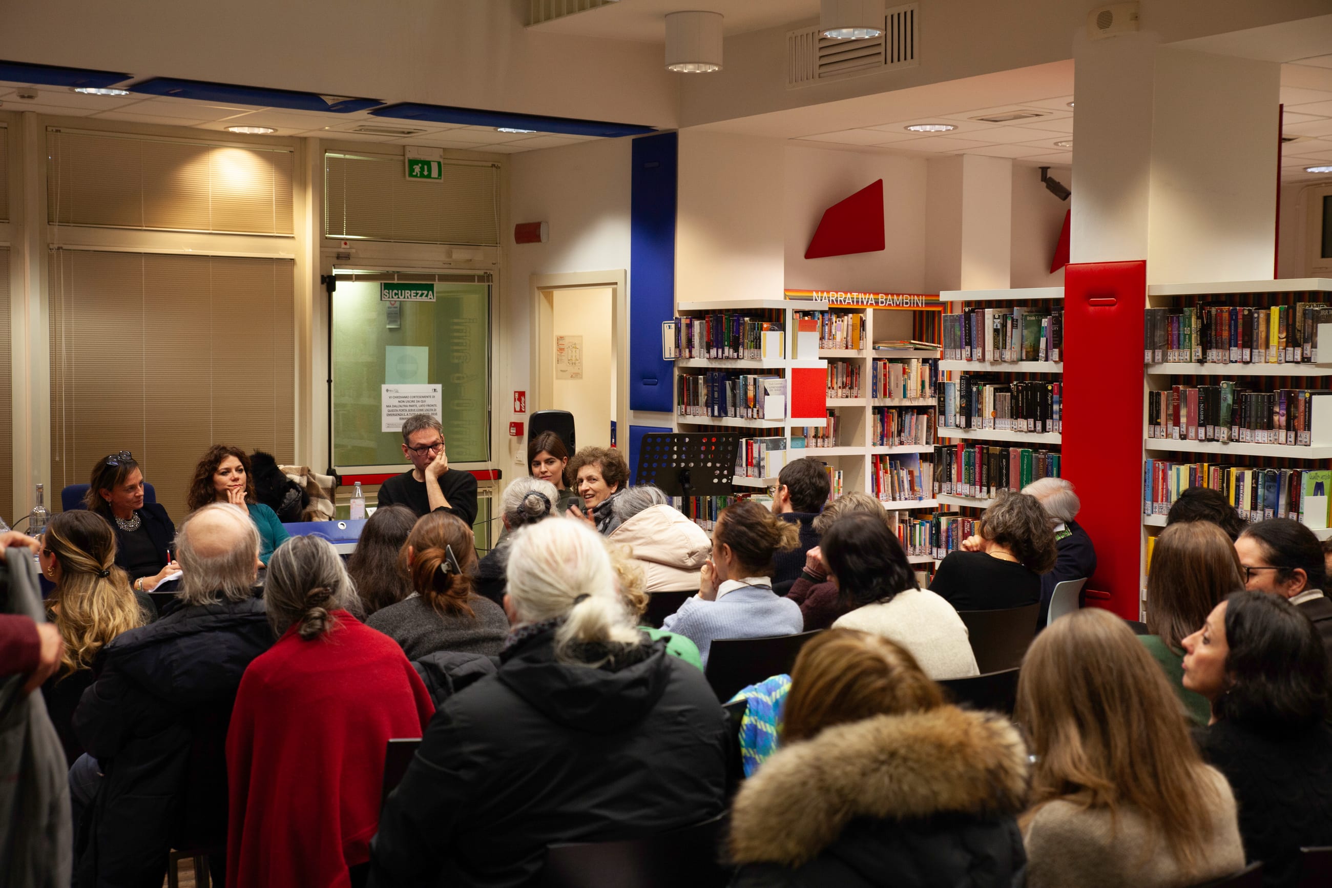 Pubblico seduto in una biblioteca tra scaffali di libri, visto da dietro