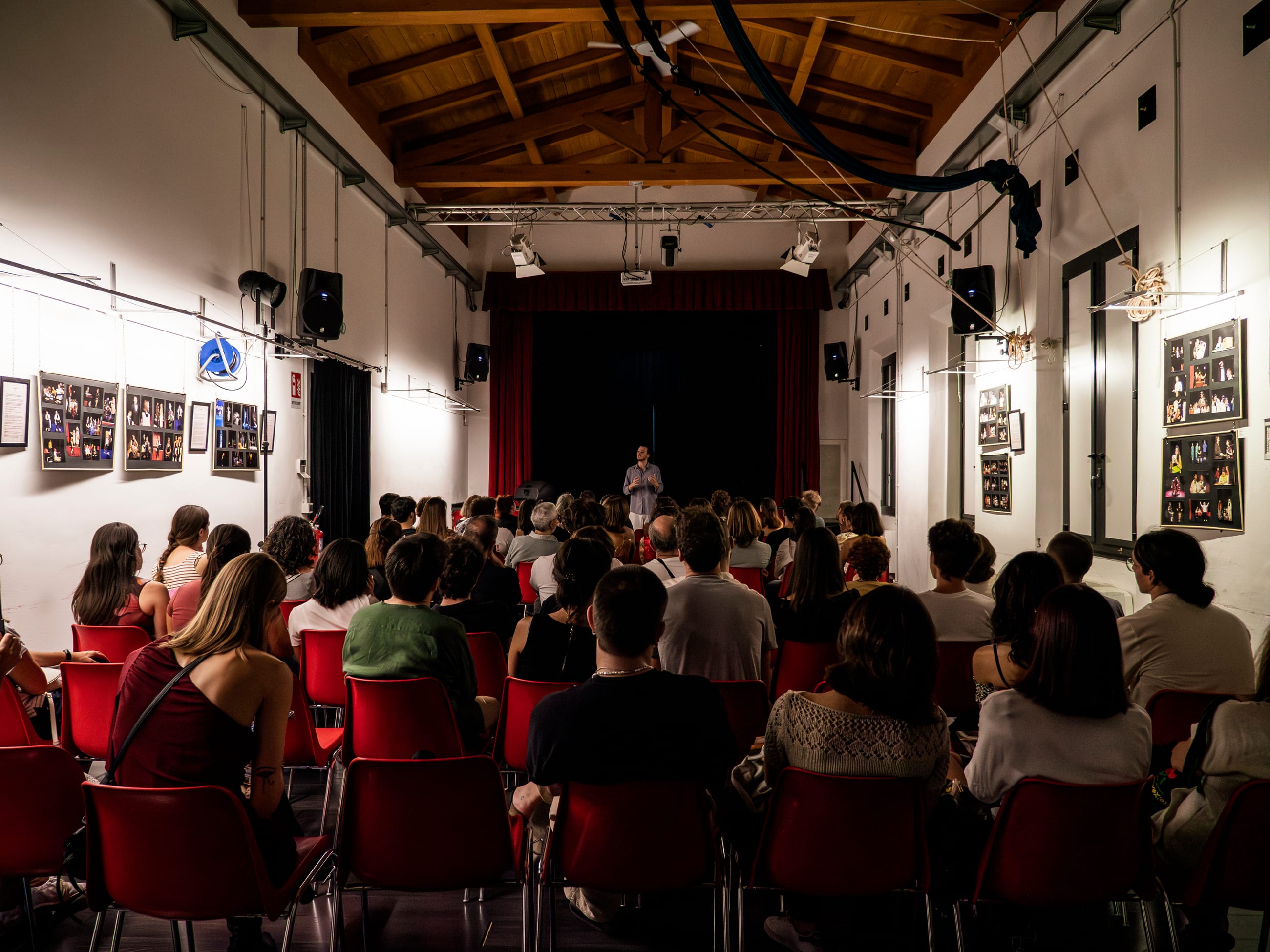 Sala con travi in legno e pubblico seduto su sedie rosse durante un incontro