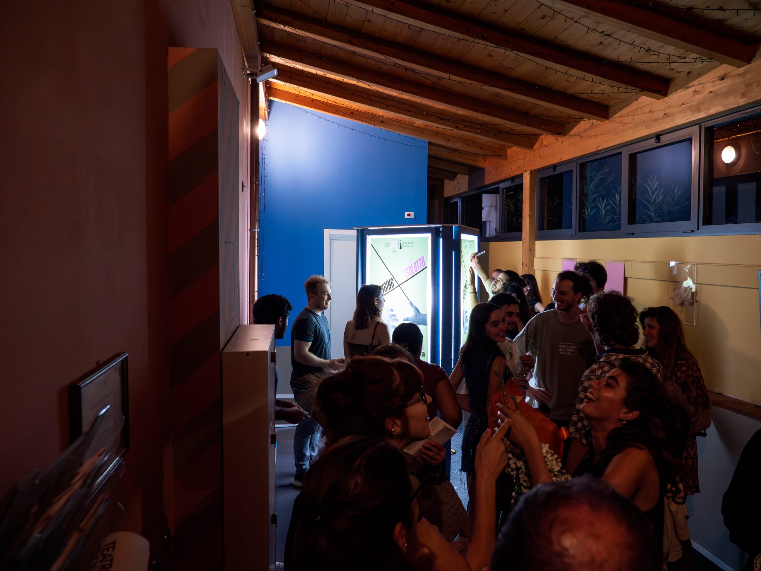 Persone in piedi in uno spazio affollato con soffitto in legno e un pannello luminoso