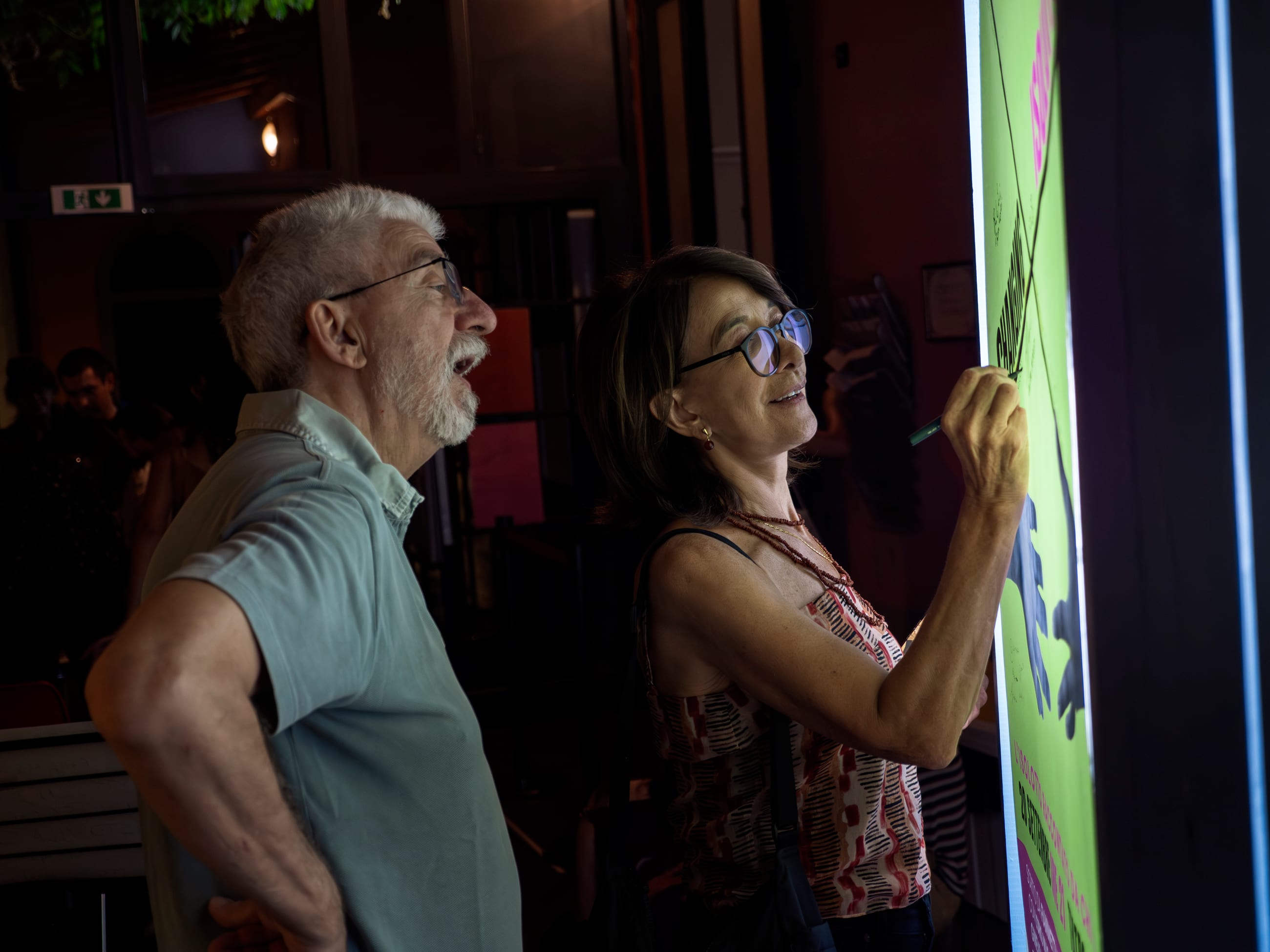 Due persone davanti a un poster luminoso verde, una delle due scrive con una matita