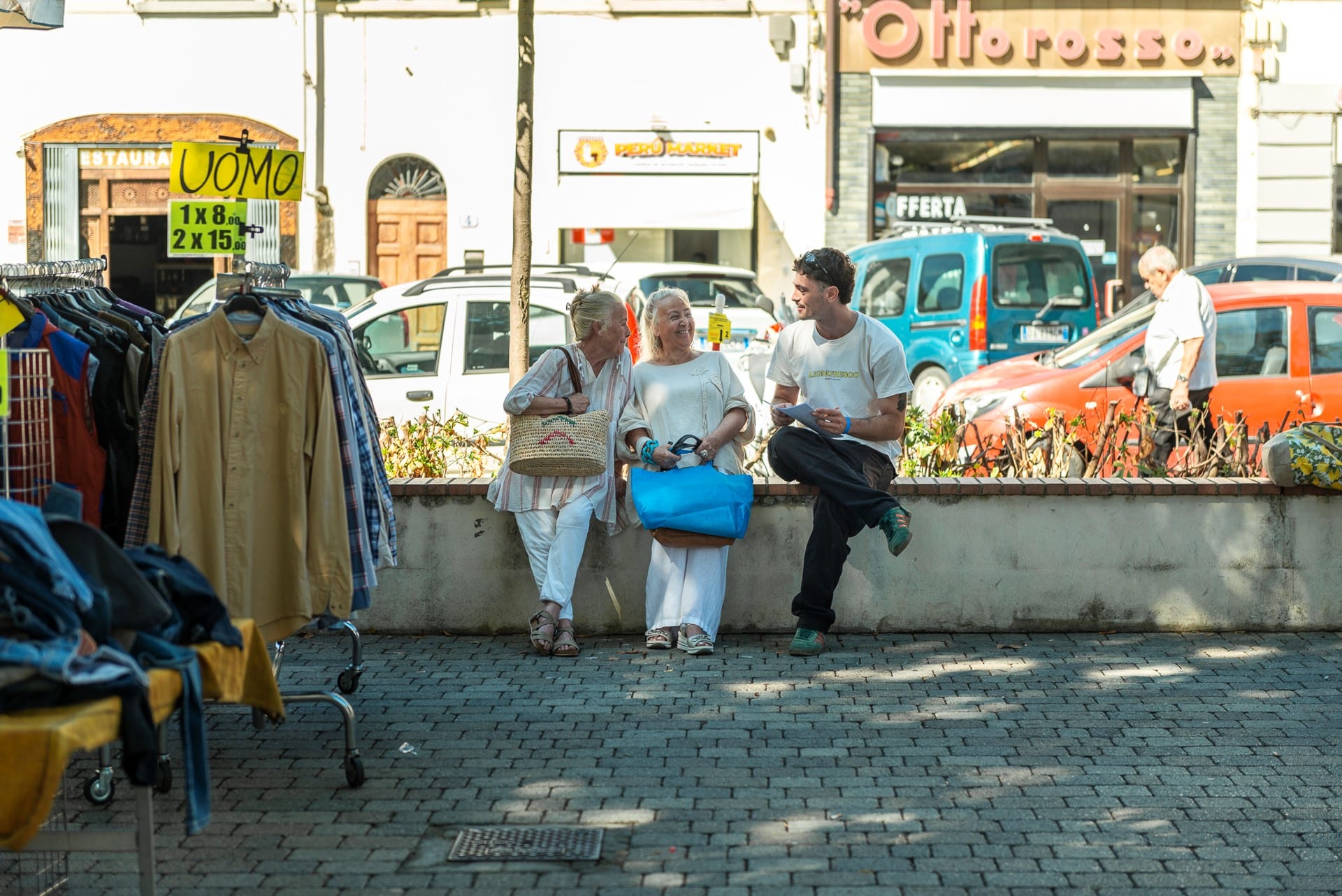 Persone sedute su un muretto in una piazza, accanto a un banco di abiti del mercato