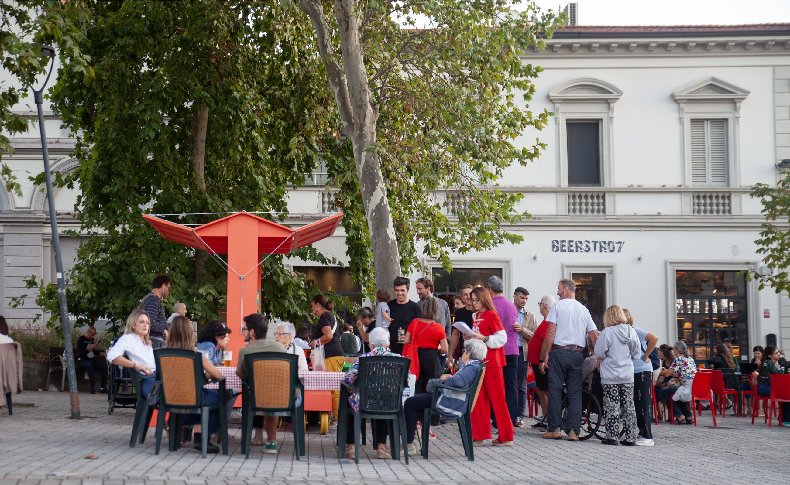 Persone sedute ai tavoli in una piazza alberata, attorno alla struttura arancione