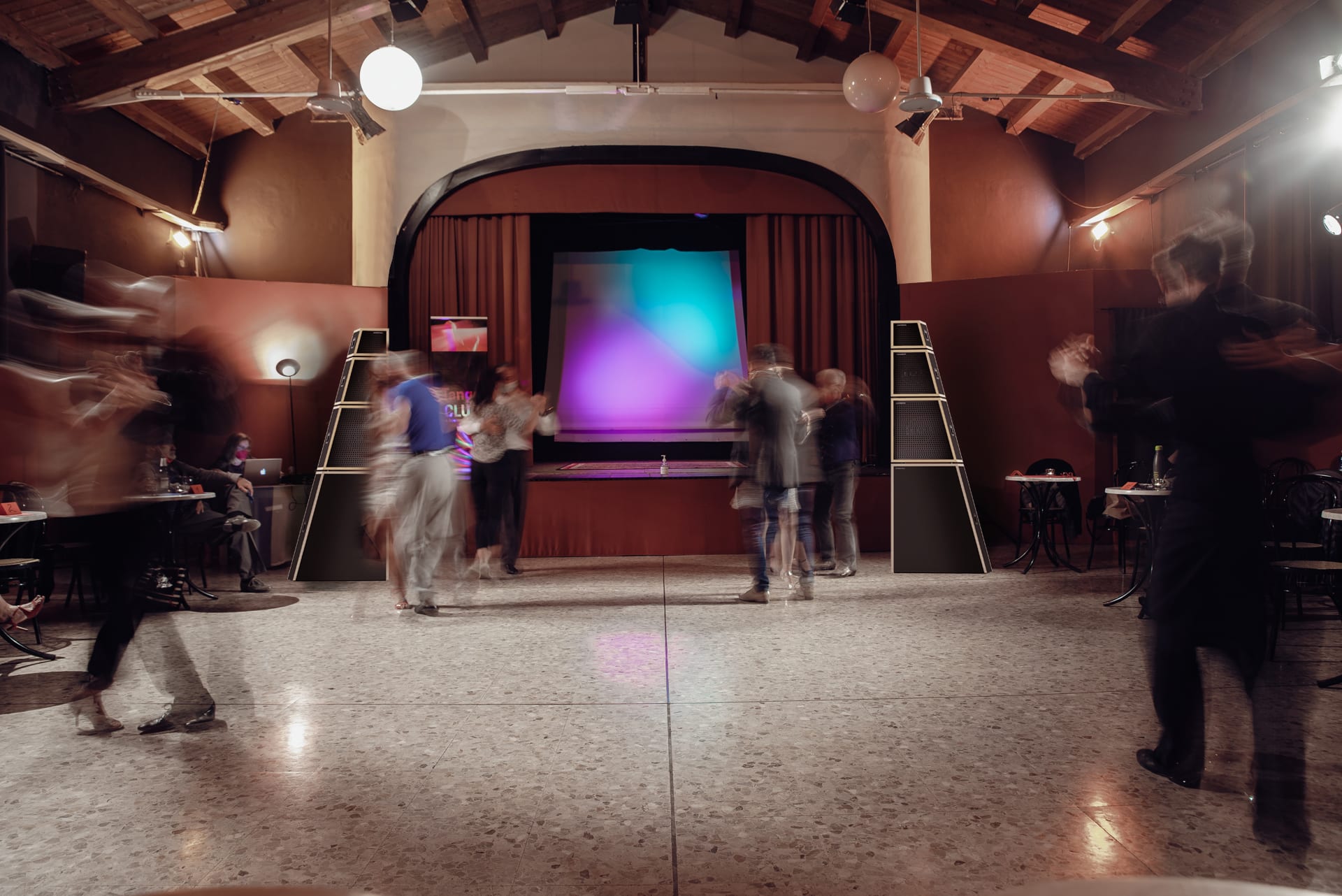 Persone in movimento in una sala con palco, tra due torri di casse audio