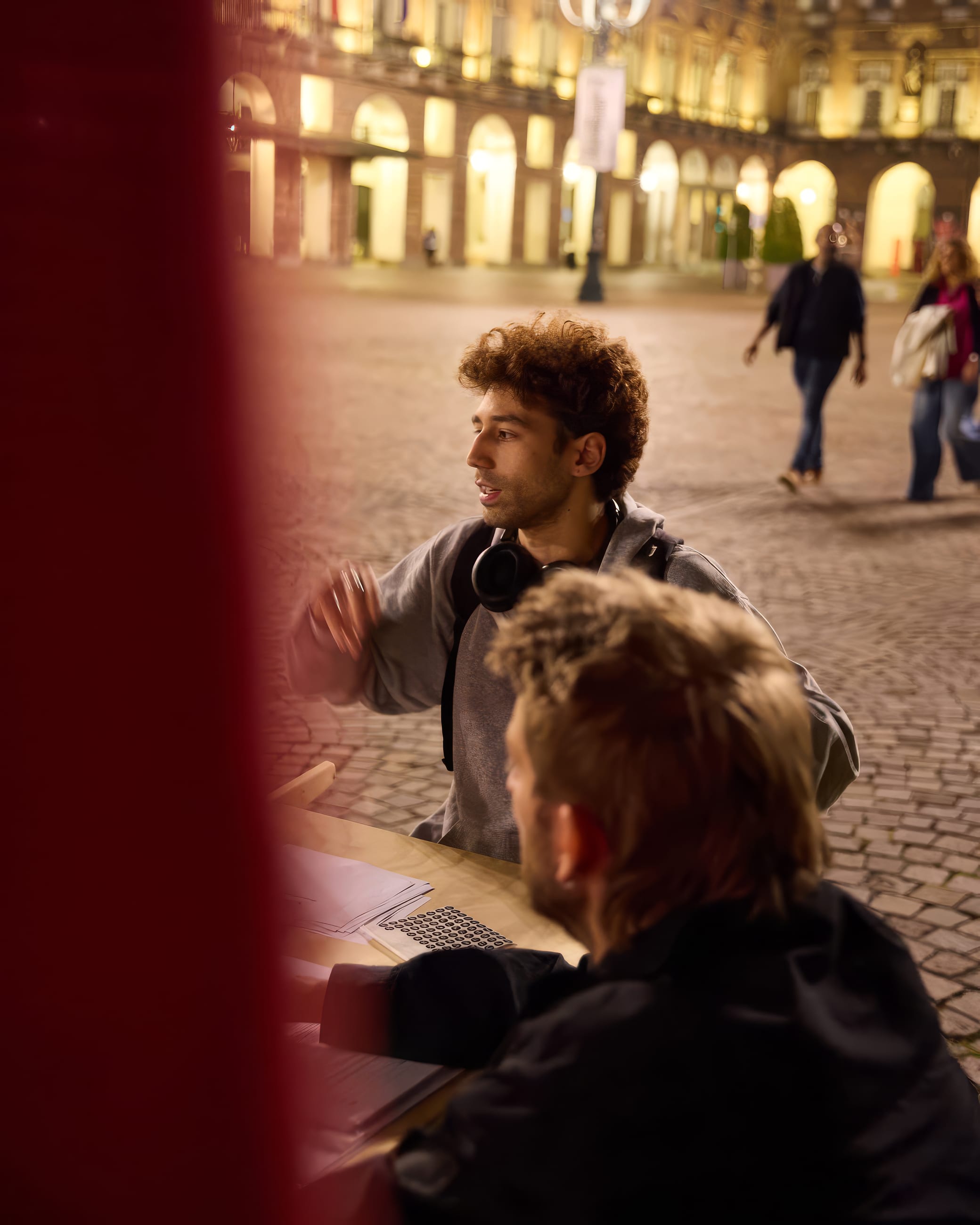 Due persone conversano a un tavolo di sera, in una piazza porticata vista oltre una tenda rossa