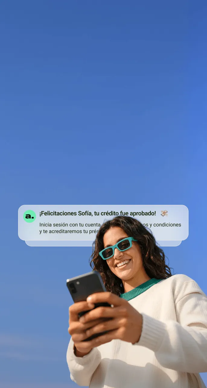 Mujer sonriente con gafas de sol verdes sosteniendo un teléfono móvil bajo un cielo azul, con un mensaje que confirma la aprobación de su crédito.