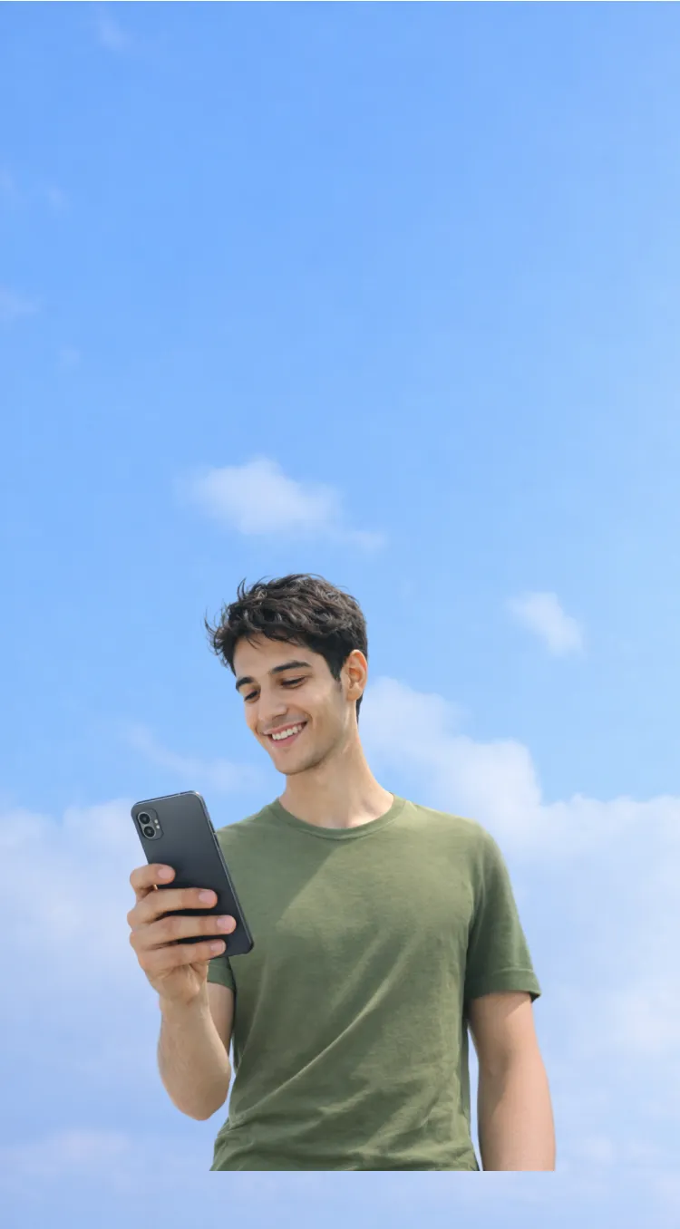 Joven sonriente vestido con camiseta verde mirando un teléfono móvil contra un cielo azul con nubes.