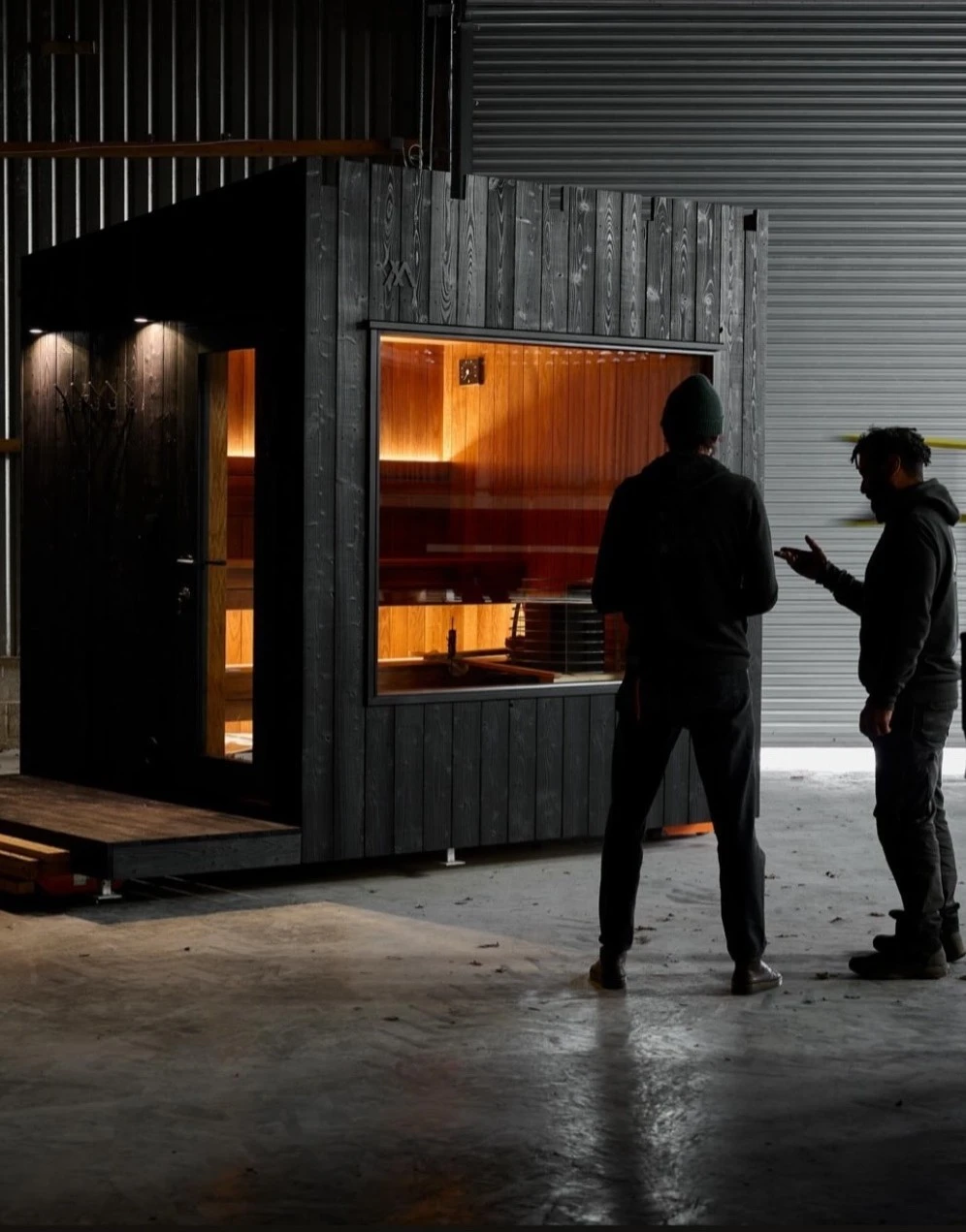 Two people standing in front of a small wooden room with dark exterior and a large glass window showing a warmly lit interior with wood paneling, inside a spacious industrial building with concrete floor and metal walls.