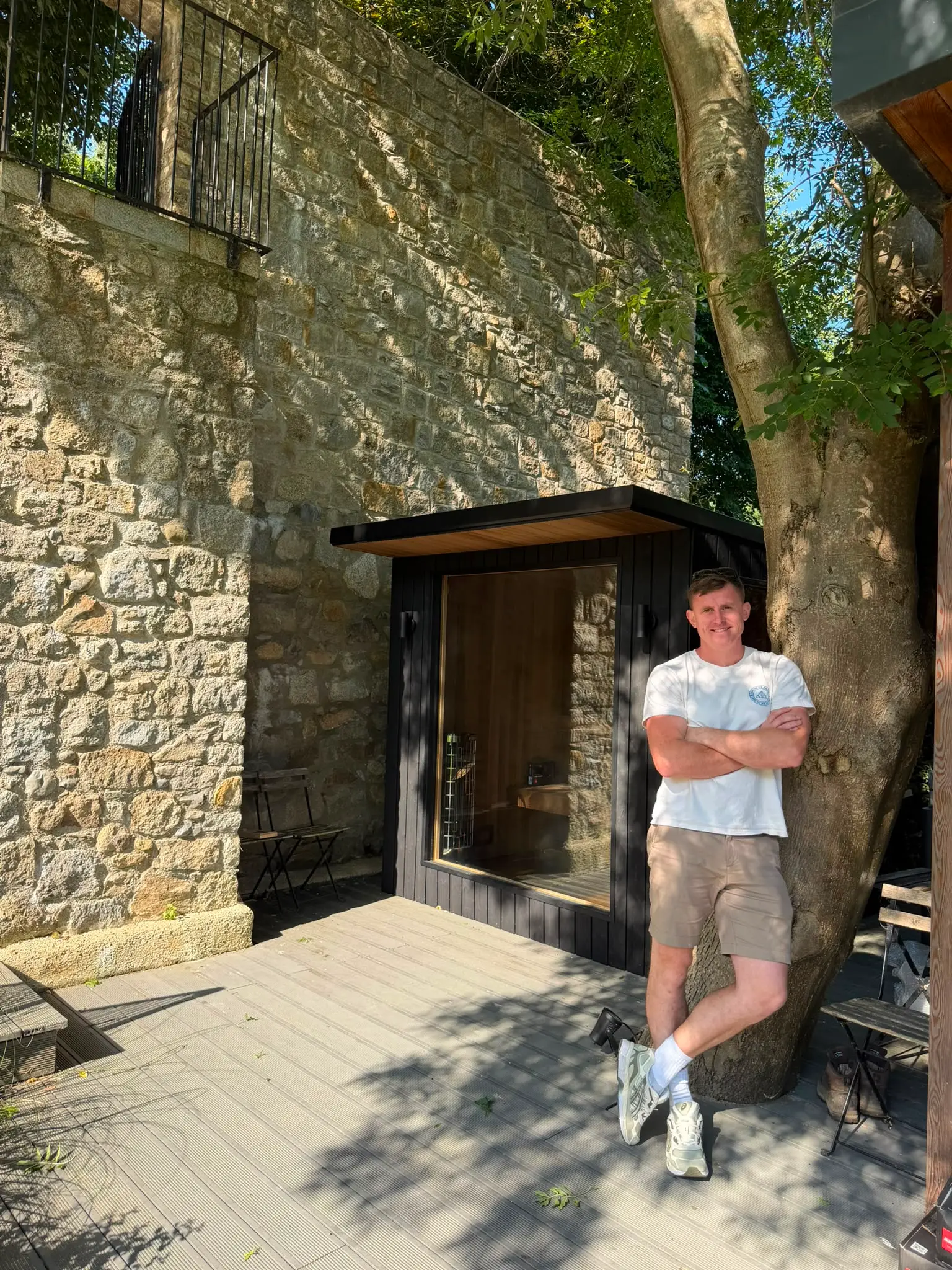 Man wearing white t-shirt, khaki shorts, and sneakers leaning against a large tree on a wooden deck next to a small black cabin with a large window, beside a stone wall and metal chair.