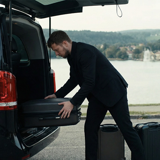 Ein Mann im schwarzen Anzug lädt einen schwarzen Koffer in den Kofferraum eines Fahrzeugs mit Seeblick. KHAN Fahrdienst Konstanz und Überlingen