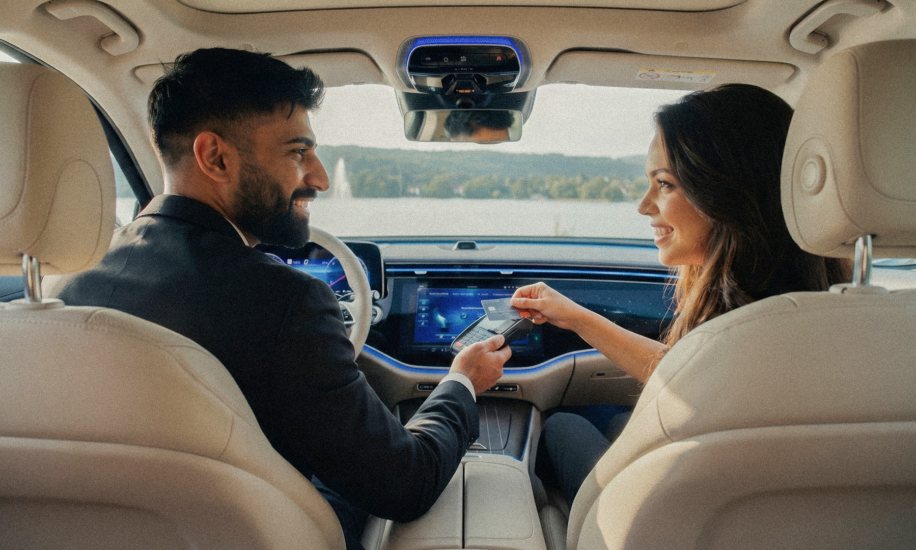 Ein Mann und eine Frau sitzen in einem Auto mit beigen Ledersitzen, die Frau hält eine Kreditkarte nah an ein mobiles Kartenlesegerät, während sie sich anlächeln. KHAN Fahrdienst Konstanz und Überlingen
