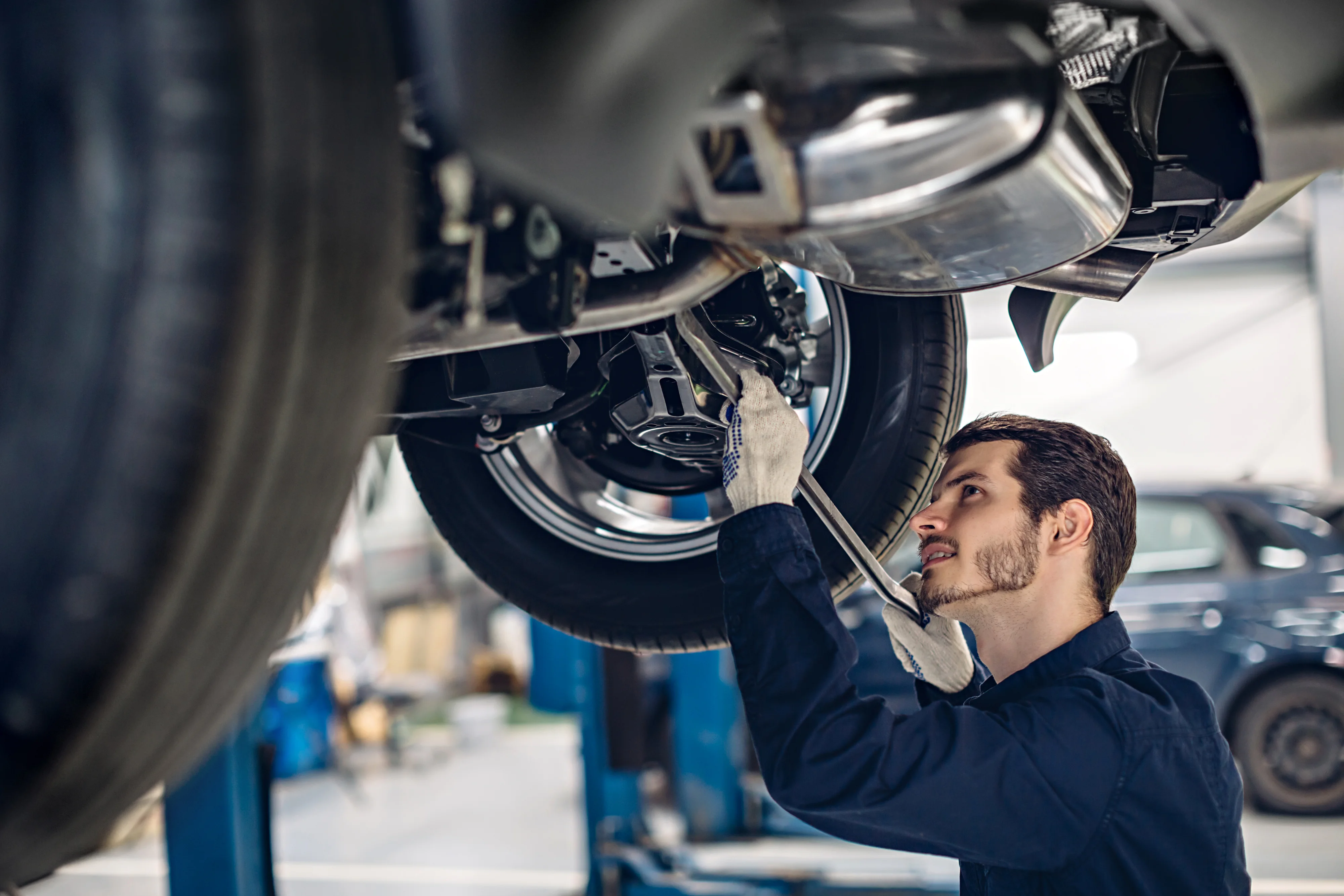 A focused mechanic in a blue uniform and gloves repairs a car's underside on a lift in a clean, bright workshop, conveying a sense of professionalism.