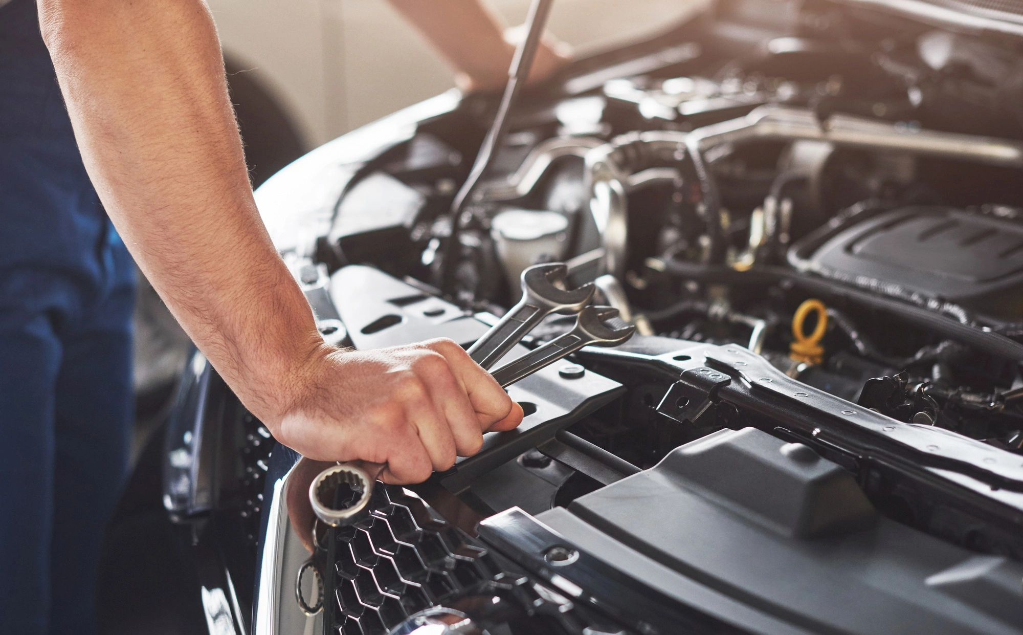 A mechanic's arm holds a wrench over an open car engine, set in a brightly lit workshop. The scene conveys focus and hands-on repair work.