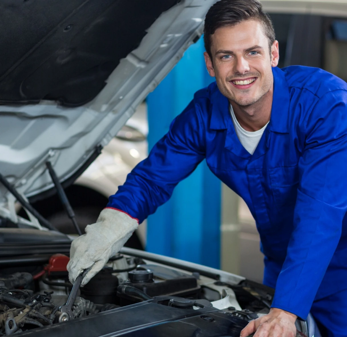 Mechanic in blue coveralls smiles while working on a car engine with the hood open. The setting is a garage, conveying a positive, professional tone.