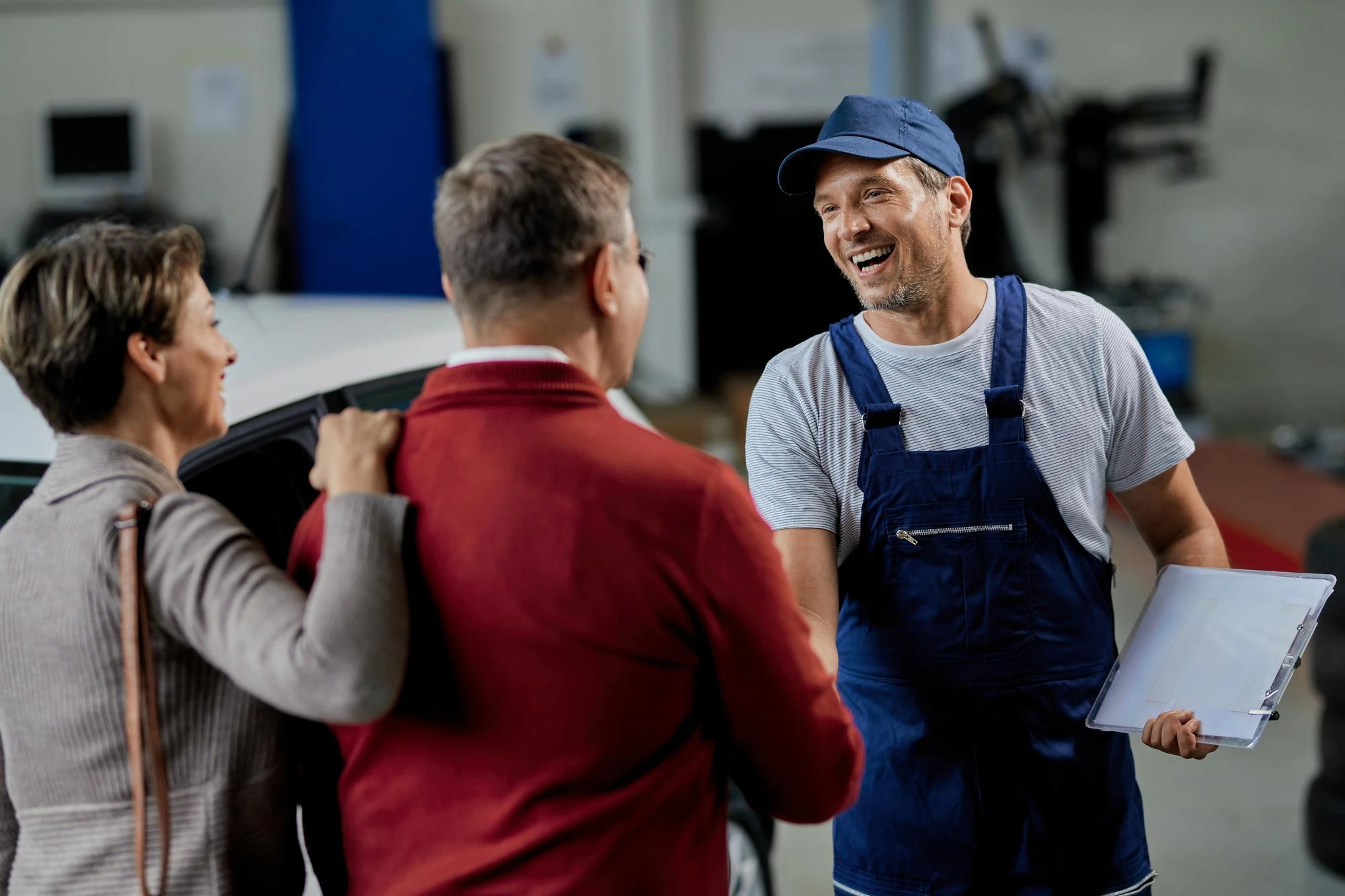 A smiling mechanic in blue overalls and cap shakes hands with a couple, conveying friendliness. They stand in a bright auto repair shop, exuding trust.