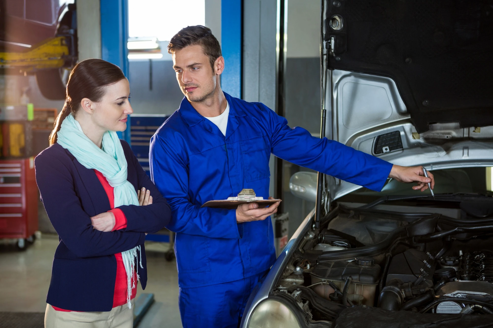 A mechanic in a blue jumpsuit shows a woman an open car hood in a garage. He holds a clipboard, explaining repairs, as she listens attentively, arms crossed.