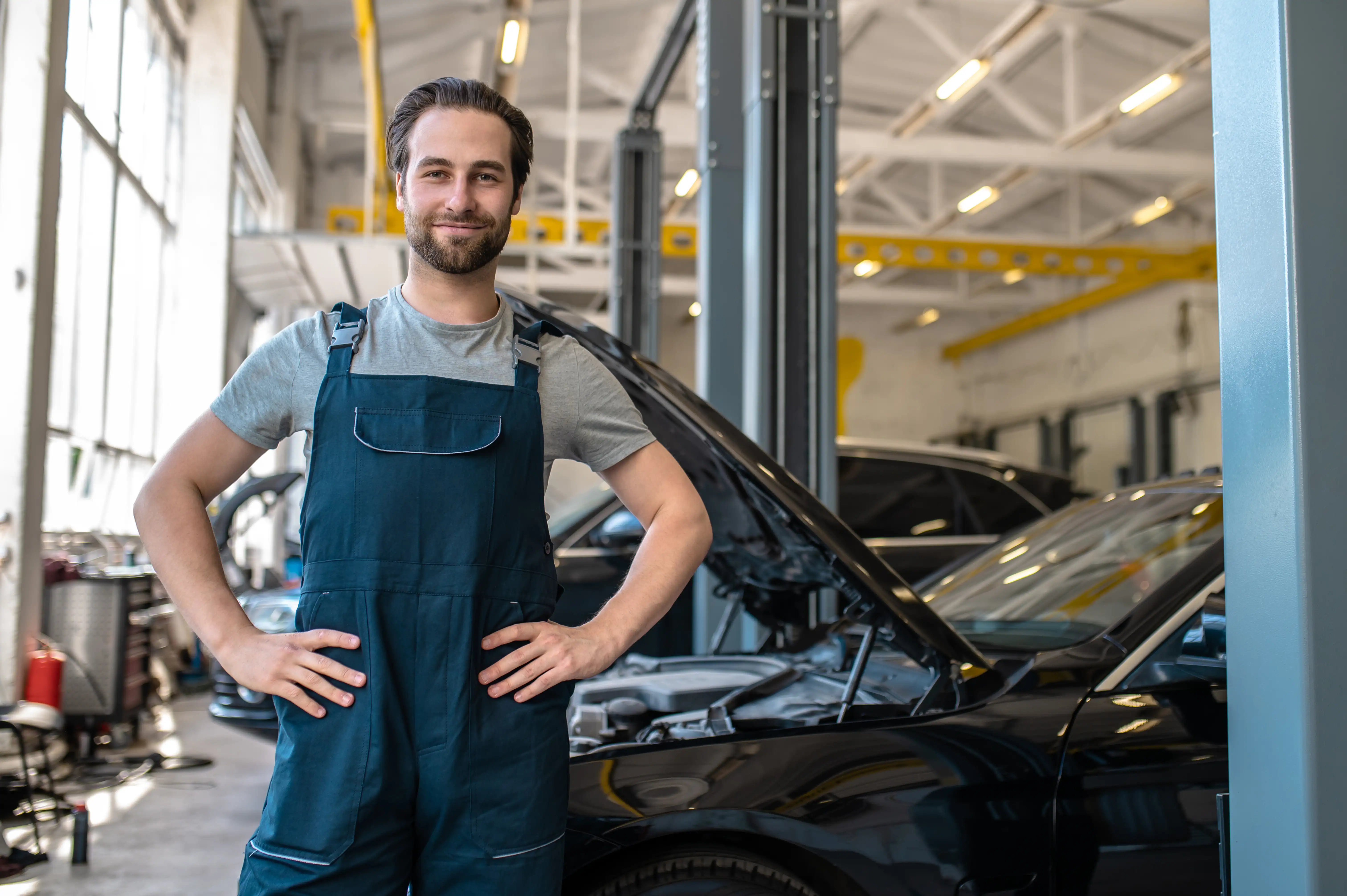 A smiling mechanic in overalls stands confidently with hands on hips in a brightly lit garage. A car with an open hood is visible in the background.