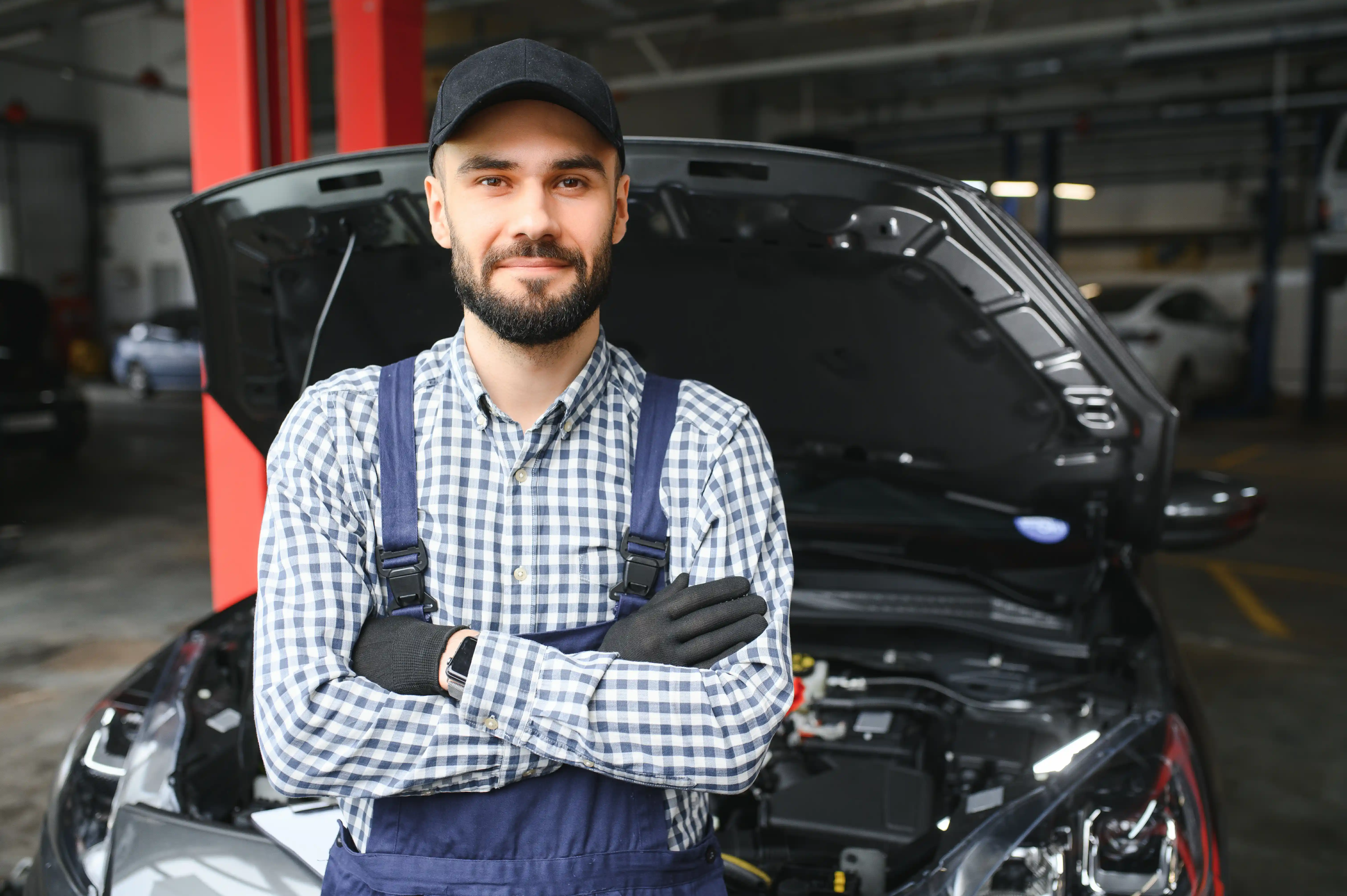 A mechanic in a plaid shirt and overalls, wearing a cap and gloves, confidently stands with crossed arms in front of a car with an open hood in a garage.
