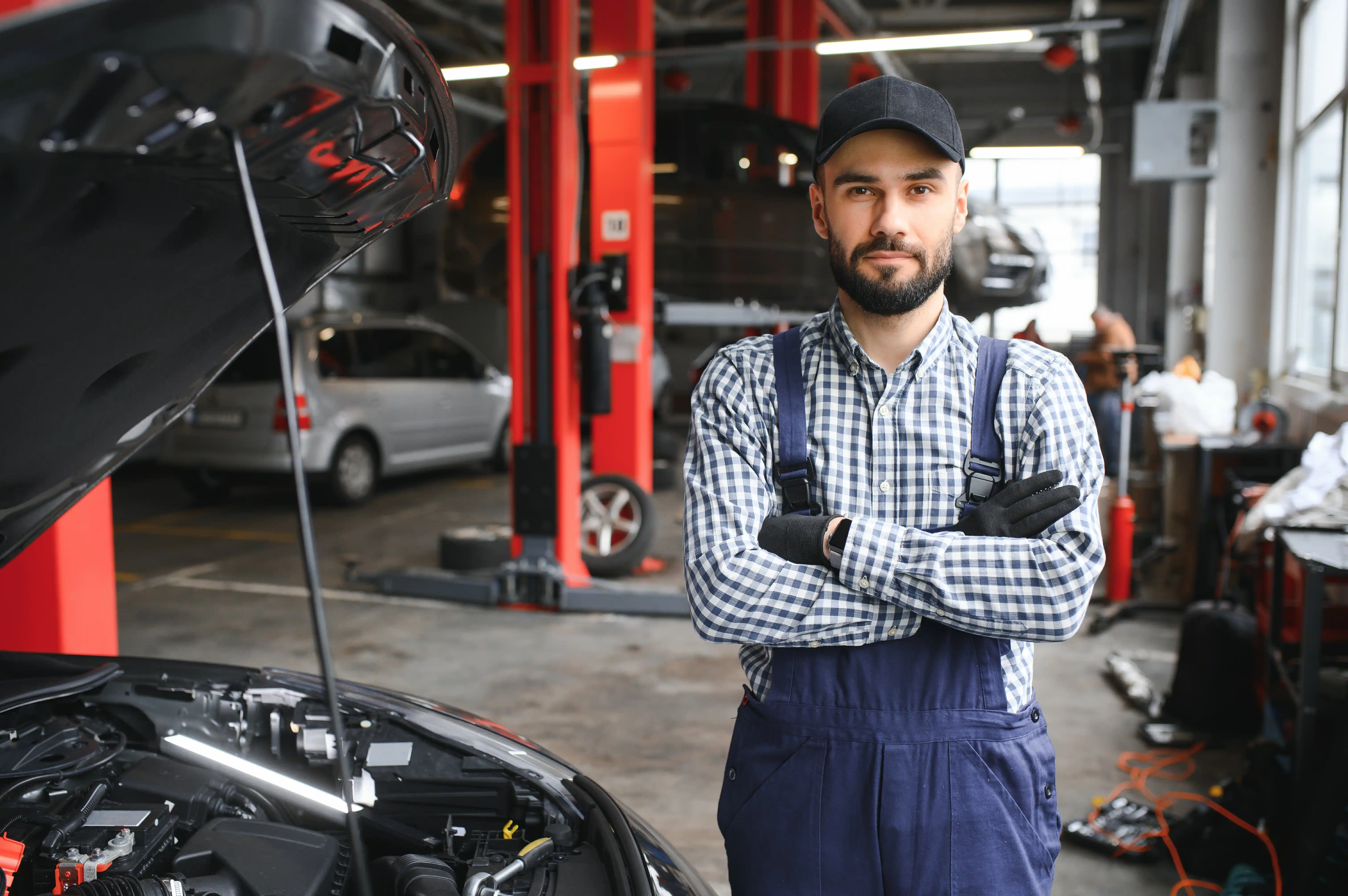 Mechanic in a cap and blue overalls stands confidently with arms crossed in an auto repair shop. Car hood is open, and vehicles are in the background.