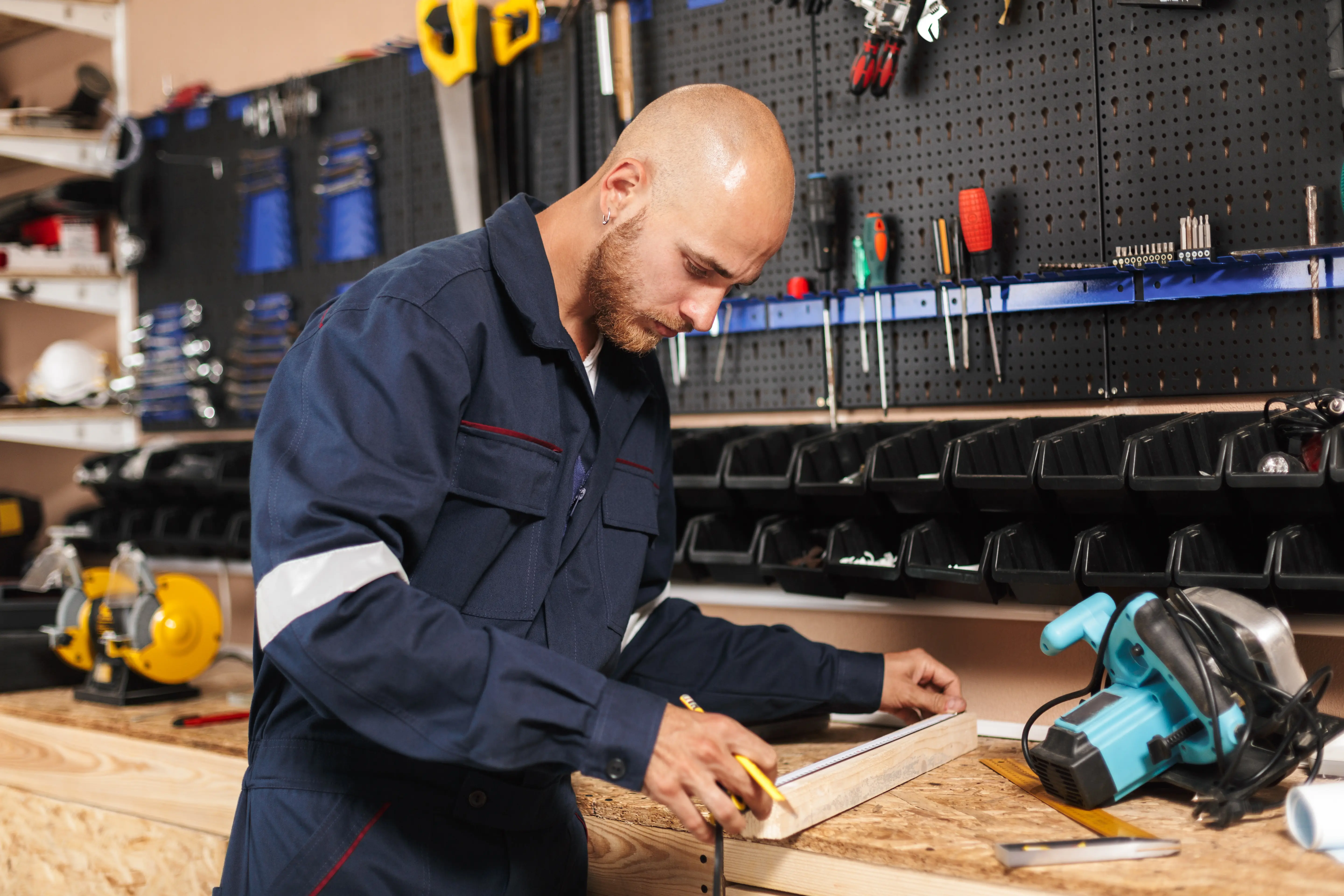Mechanic in blue overalls and cap uses a laptop in a garage, with cars on lifts in the background. He appears focused and engaged.