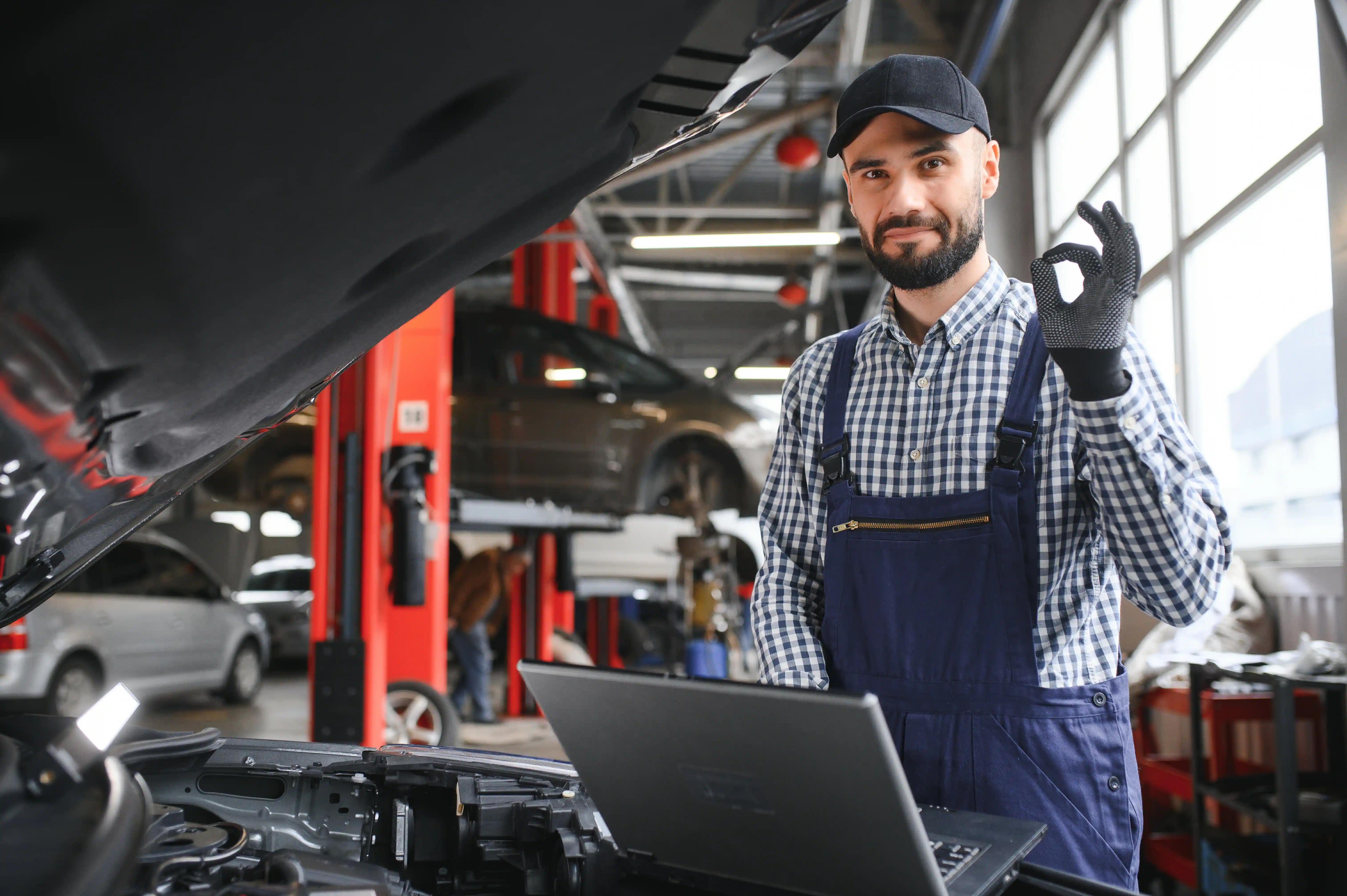 Mechanic in overalls and a cap gives an "OK" sign beside a car hood, with a laptop on the engine. Workshop with lifts and cars in the background.