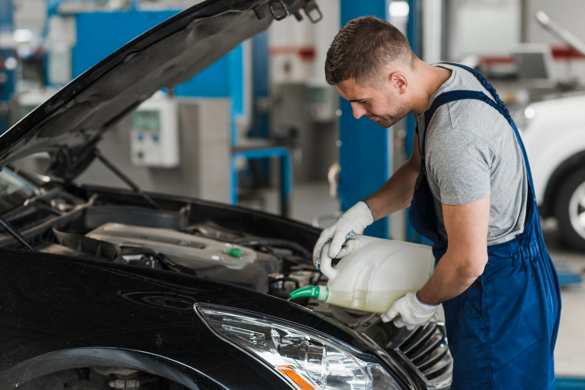 A mechanic in blue overalls and gloves adds fluid to a car engine in a garage. The car's hood is open, and the setting is a bright, industrial workshop.