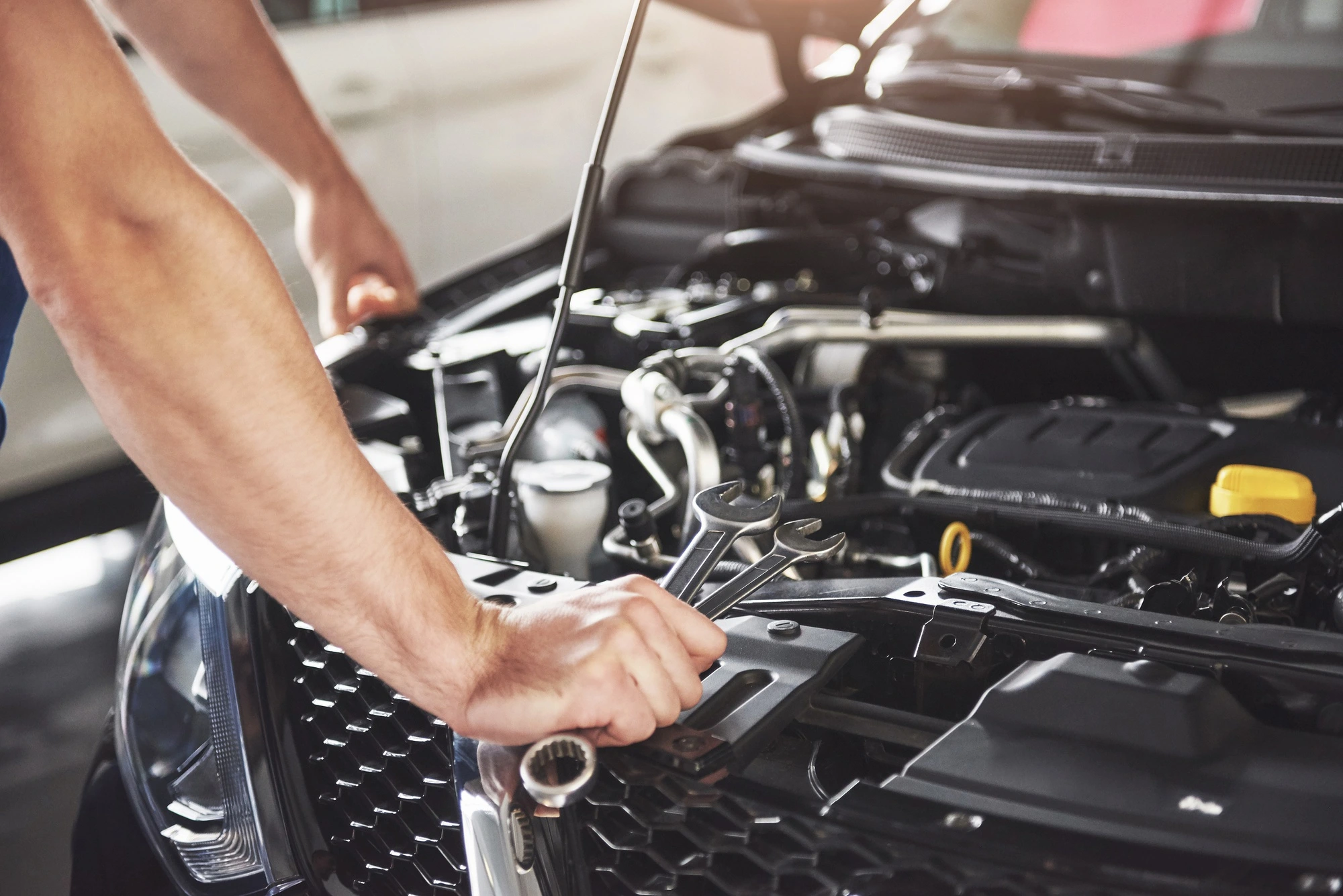 Mechanic working on a car engine, holding wrenches. The hood is open, revealing engine components. The scene conveys focus and technical skill.