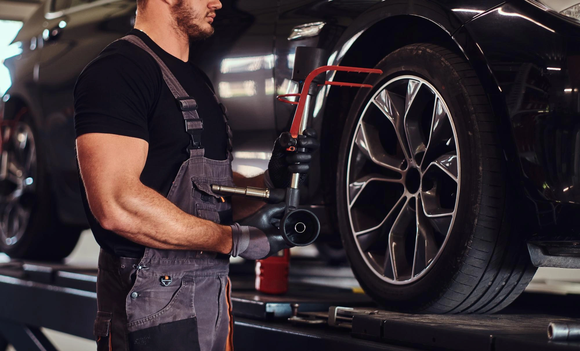 Mechanic in black shirt and overalls using a tool to repair a black car on a lift. He is focused on a tire, with tools around him, conveying diligence.