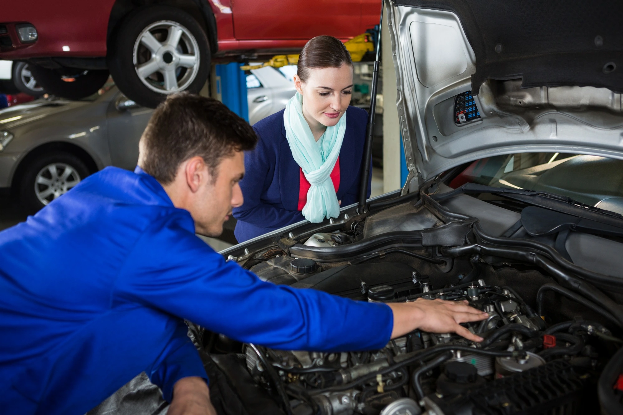 A mechanic in blue overalls explains car engine details to a woman in a scarf at an auto shop. Both are focused and attentive, surrounded by cars.