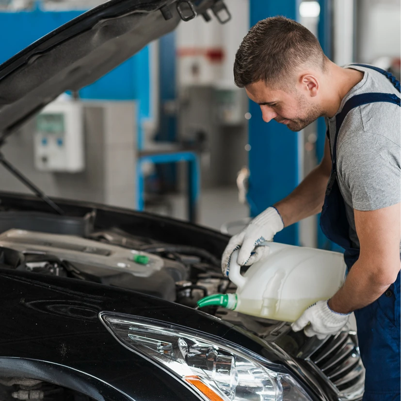 Mechanic in blue overalls and gloves fills a car radiator with coolant from a white container in a bright garage, displaying focus and expertise.