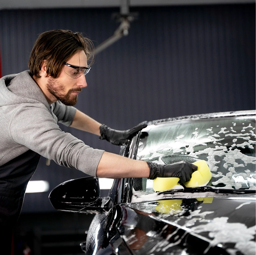 A focused individual wearing safety glasses and gloves cleans a car's windshield using a yellow sponge. The scene conveys diligence and attention to detail.