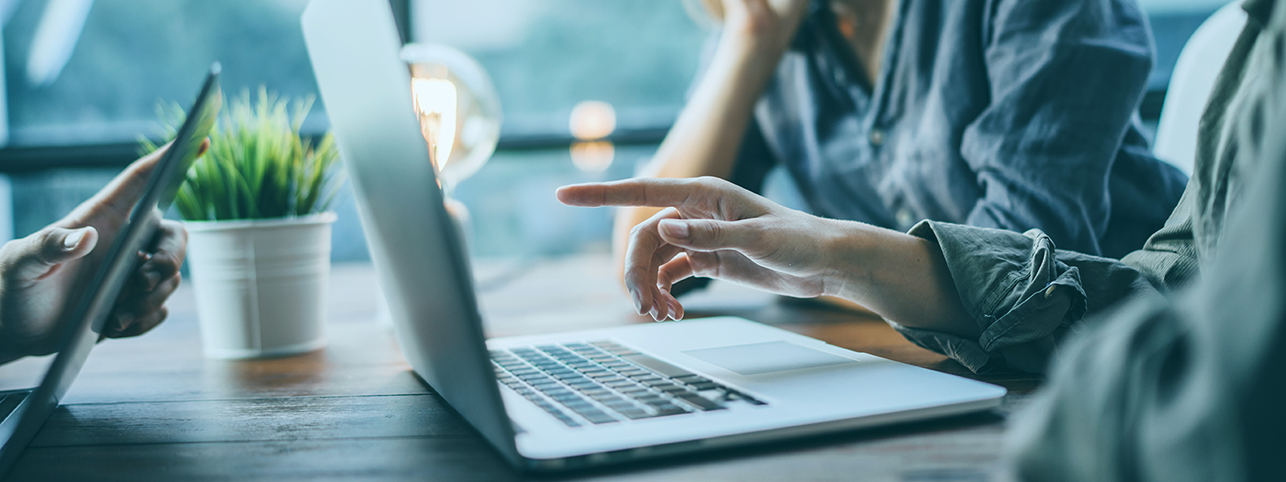 To personer arbejder sammen ved et bord med en bærbar computer og et tablet, hvor den ene peger på laptopskærmen, og en potteplante står på bordet ved siden af.