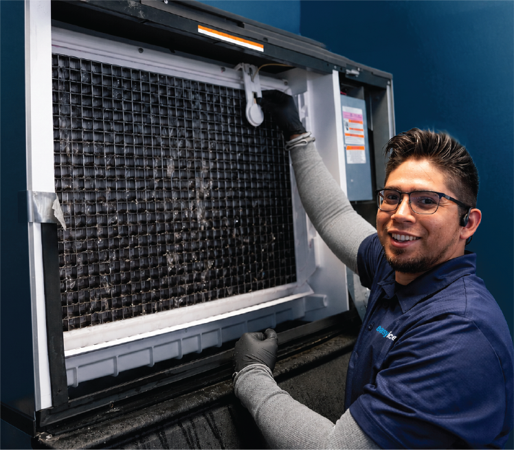 Ice machine technician smiles at camera while showing an open commercial ice machine's evaporator system with water flowing over it.