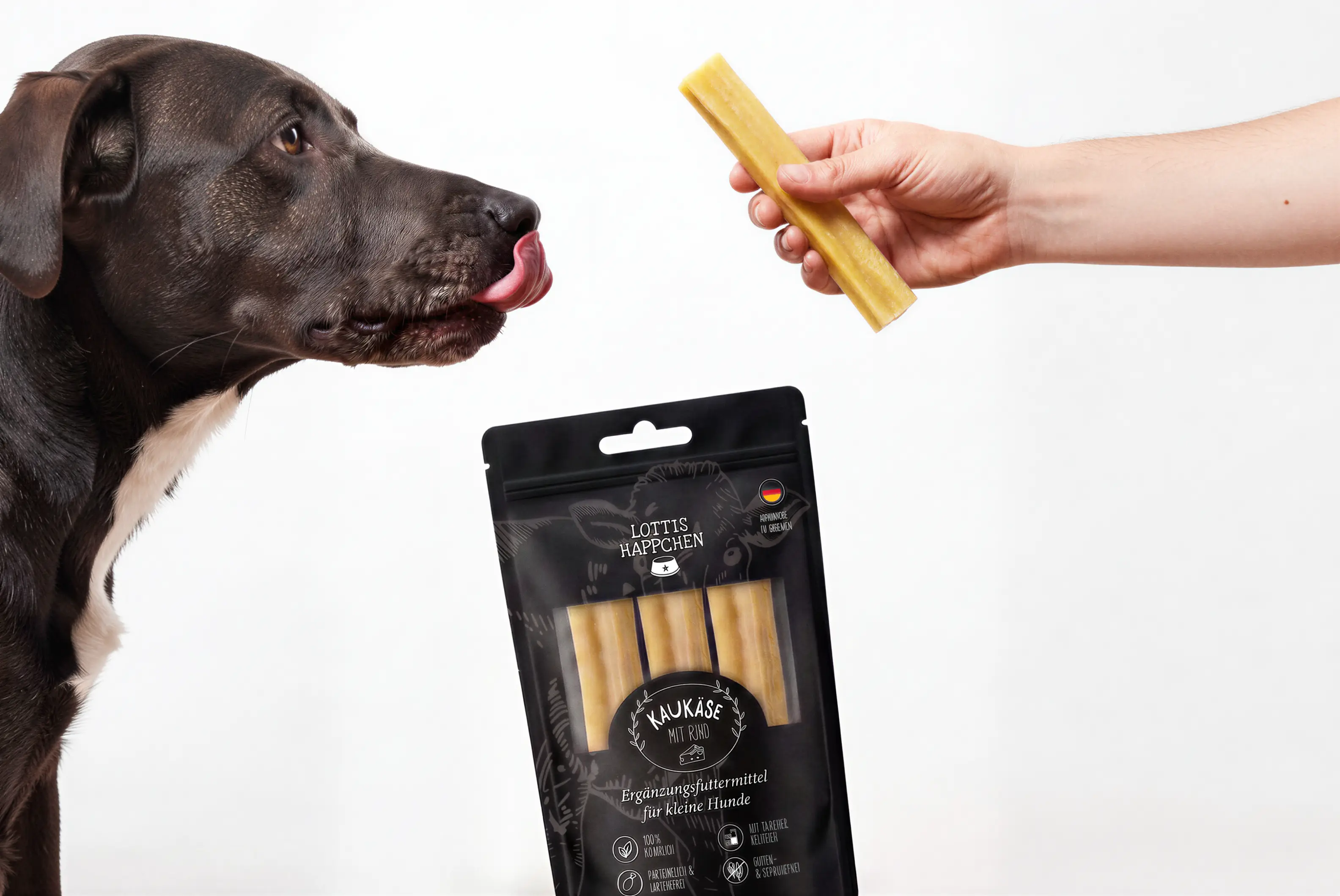 Black dog licking lips facing hand holding a yellow dog chew treat above its package.