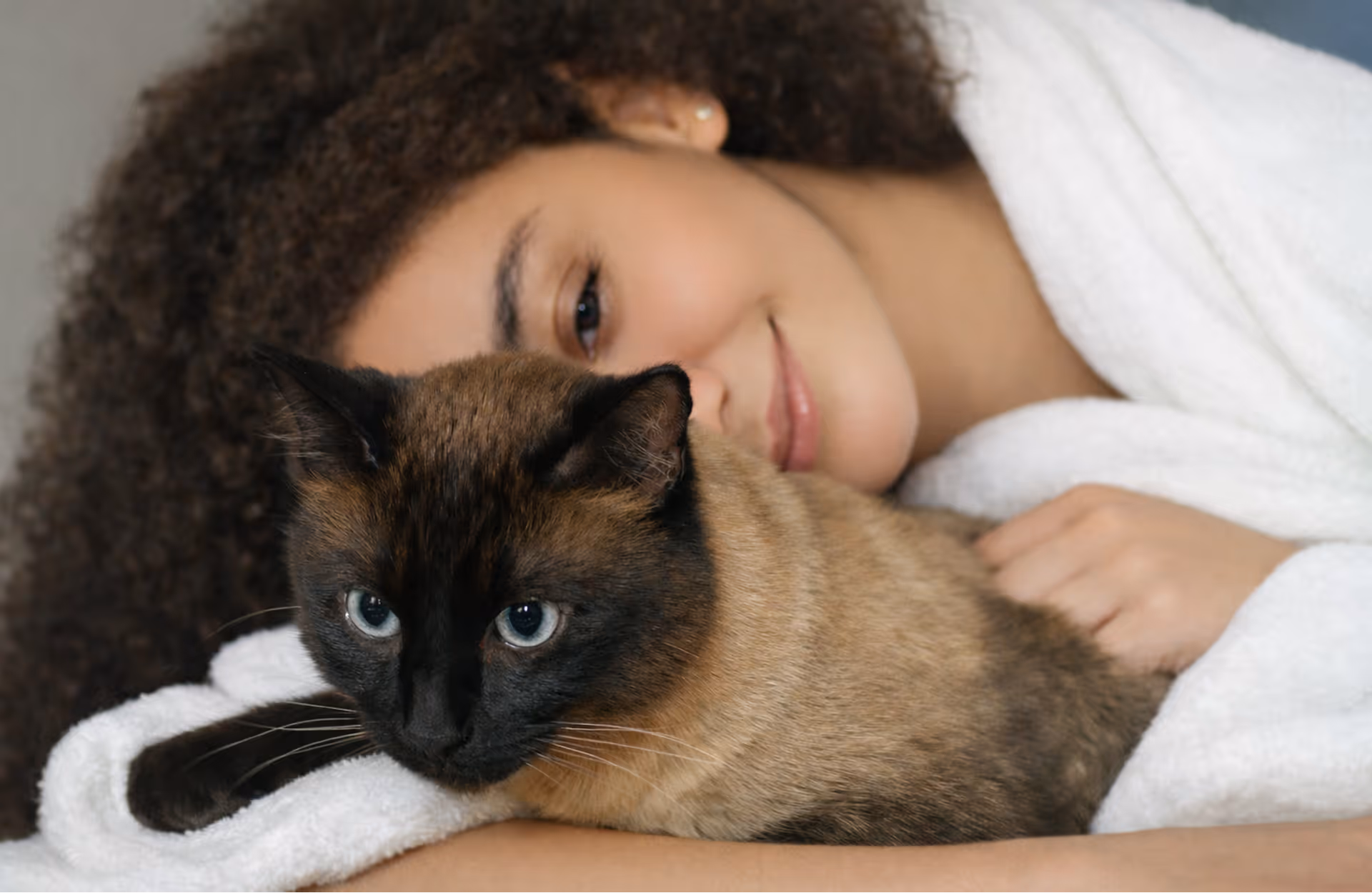 Mulher sorridente de pele clara e cabelo cacheado abraça um gato siamês com olhos azuis, ambos deitados sobre um tecido branco.