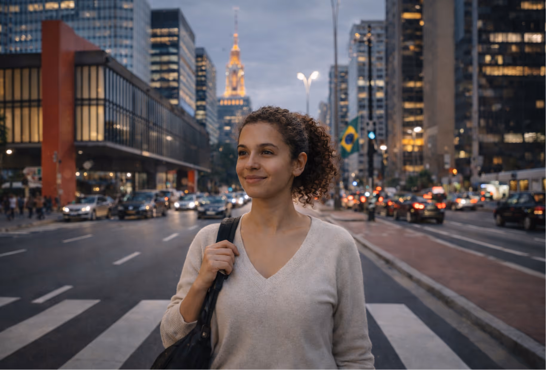 Mulher sorridente com cabelo cacheado preso andando em uma faixa de pedestres na cidade ao anoitecer, com prédios altos e carros ao redor, e uma bandeira do Brasil ao fundo.