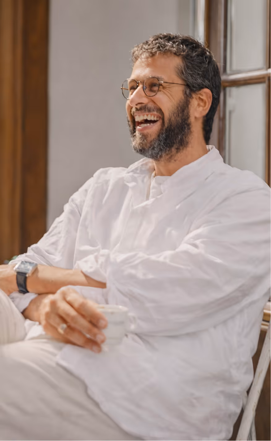Homem sorridente com barba e óculos circulares, vestido com roupa branca, sentado segurando uma xícara branca.