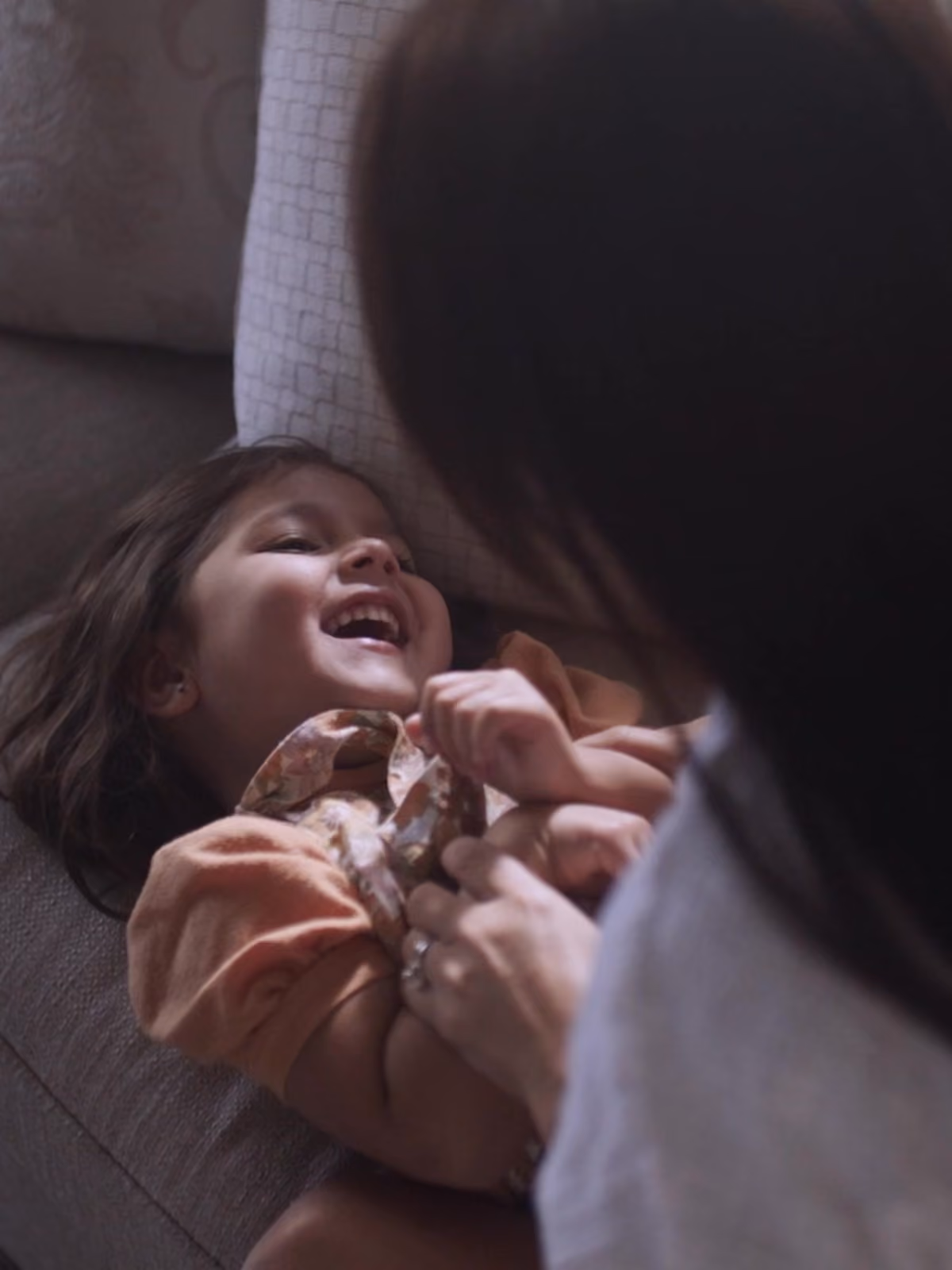 Criança sorridente deitada em um sofá sendo acariciada e abraçada por um adulto não visível completamente.