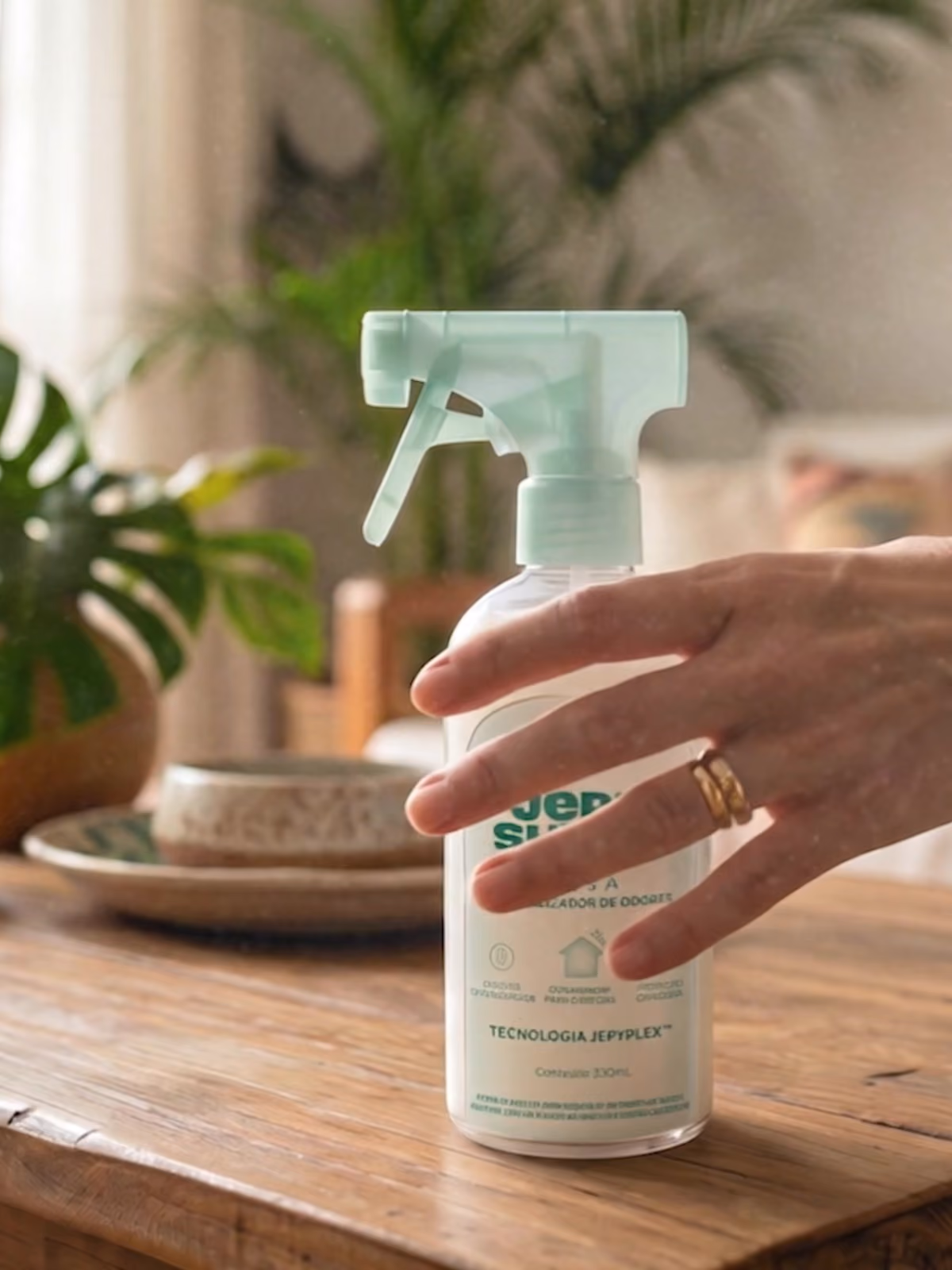 Mão segurando um borrifador branco com texto verde, sobre uma mesa de madeira, com pratos empilhados e plantas desfocadas ao fundo.
