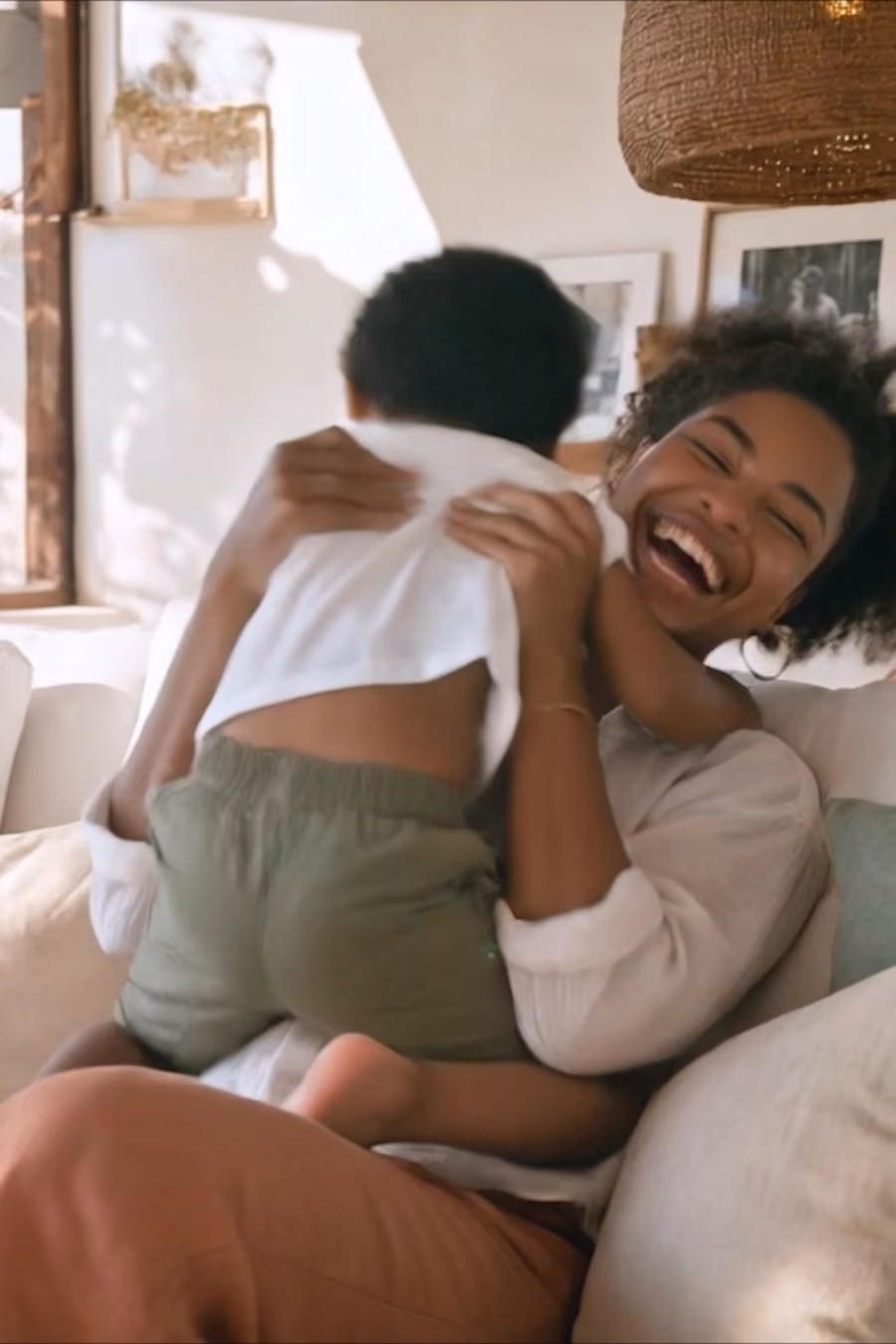 Mulher sorridente sentada em um sofá, abraçando uma criança pequena com roupa branca e calça verde, em ambiente interno com decoração acolhedora.
