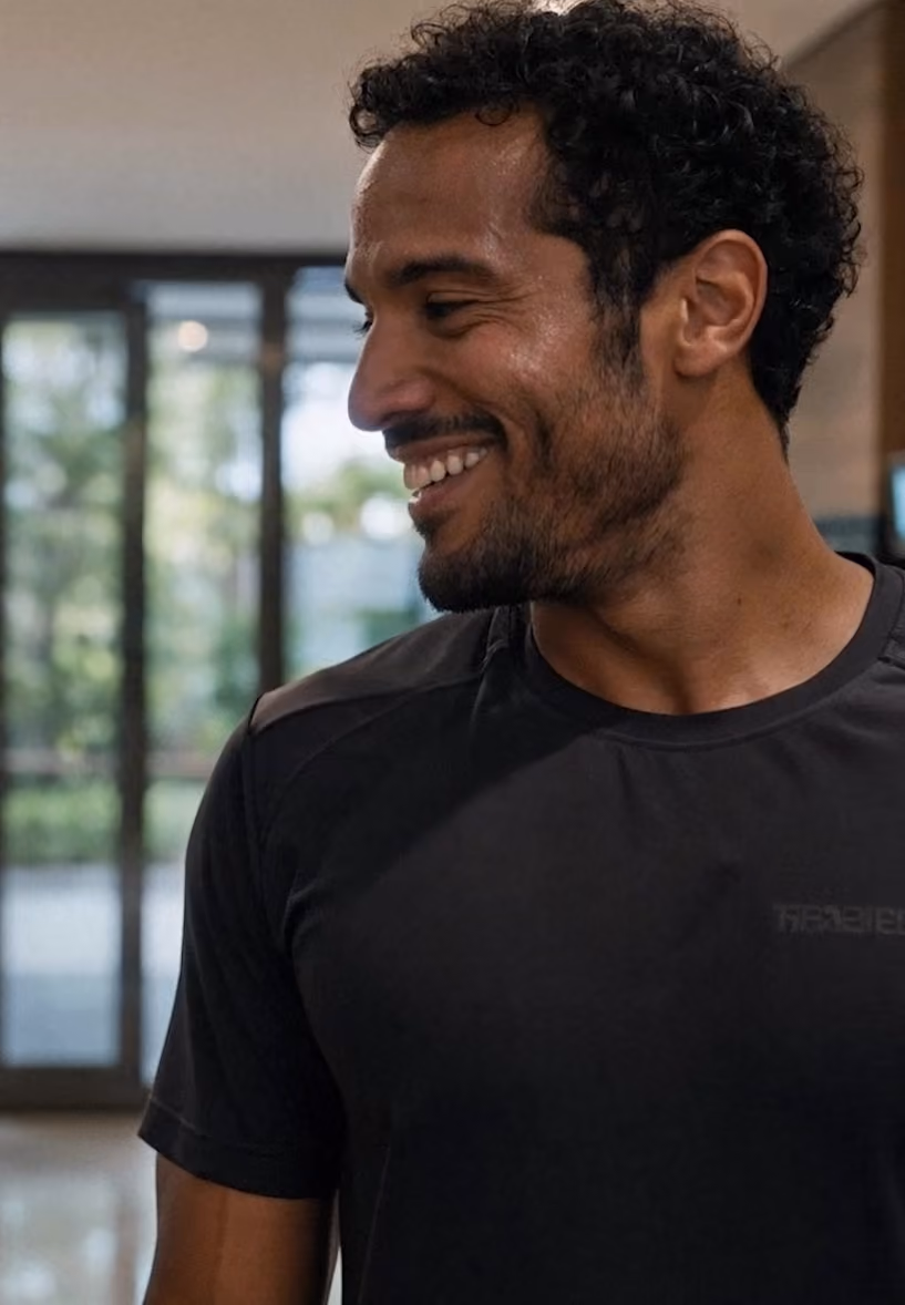 Homem sorridente com cabelos cacheados curtos e barba rala, vestindo camiseta preta, olhando para a esquerda em ambiente interno com porta de vidro ao fundo.