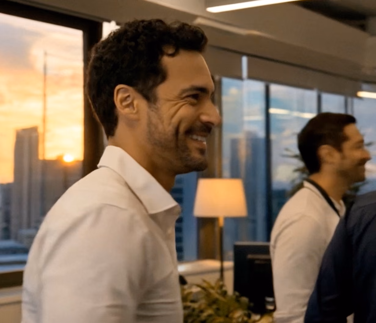 Homem sorridente de camisa branca em um escritório moderno com janelas grandes ao pôr do sol e outra pessoa desfocada ao fundo.