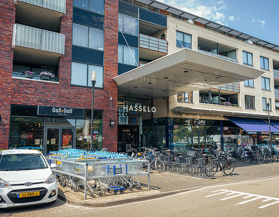 Buitenaanzicht van de hoofdingang van Winkelcentrum Hasselo met het Hasselo-logo, Gall & Gall, winkelwagens en fietsen op de parkeerplaats