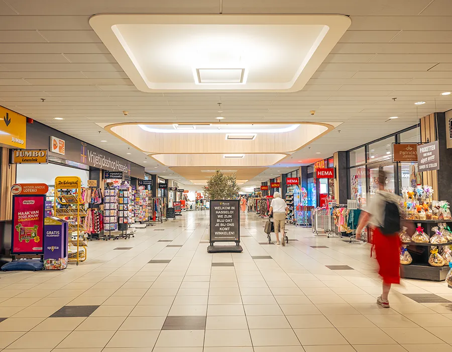 Breed interieuroverzicht van de winkelstraat in Winkelcentrum Hasselo met winkels als Jumbo, HEMA en Vrijetijdsshop Hasselo en bezoekers op de voorgrond